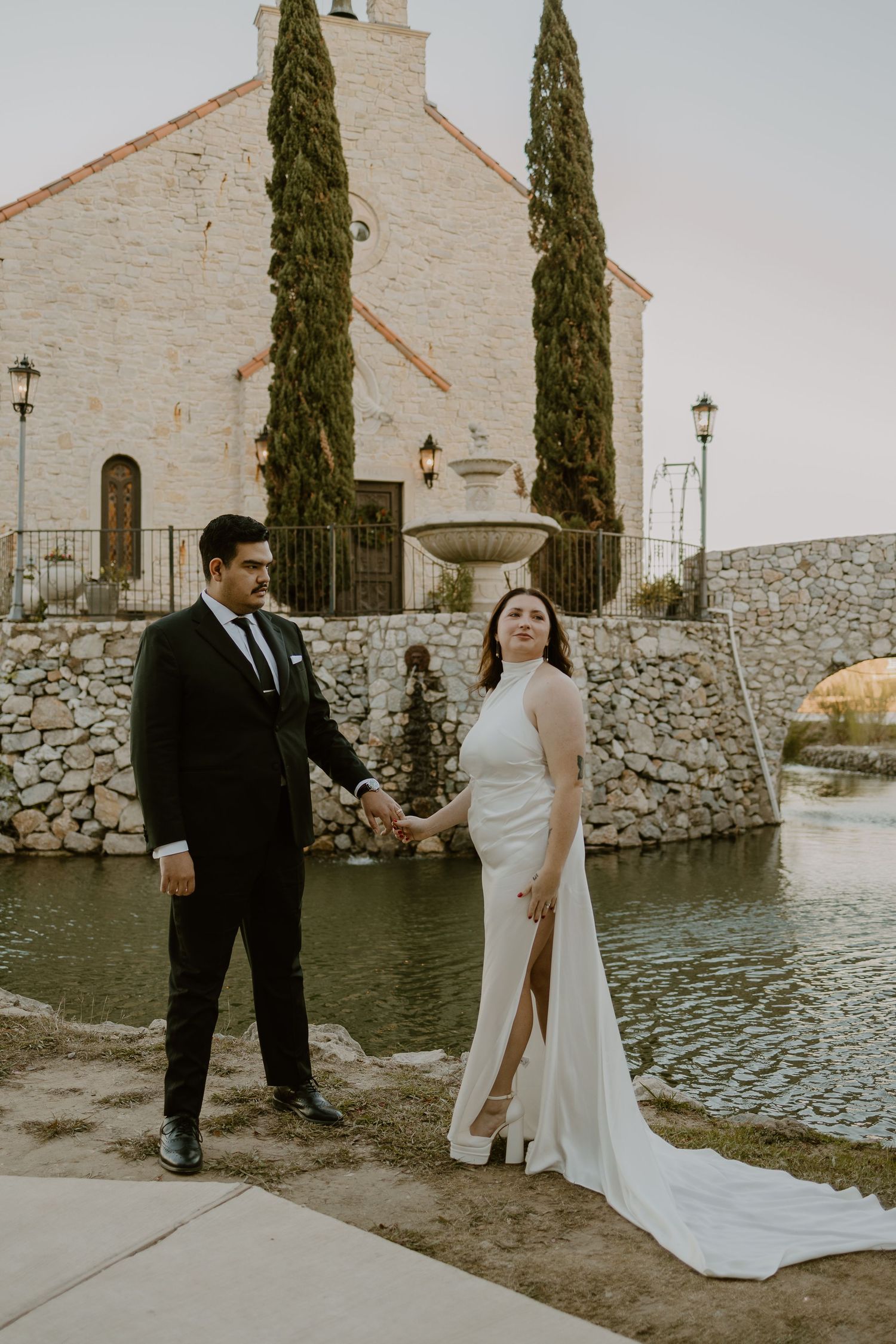 A wedding couple poses by a Mediterranean-style church with cypress trees and water feature.