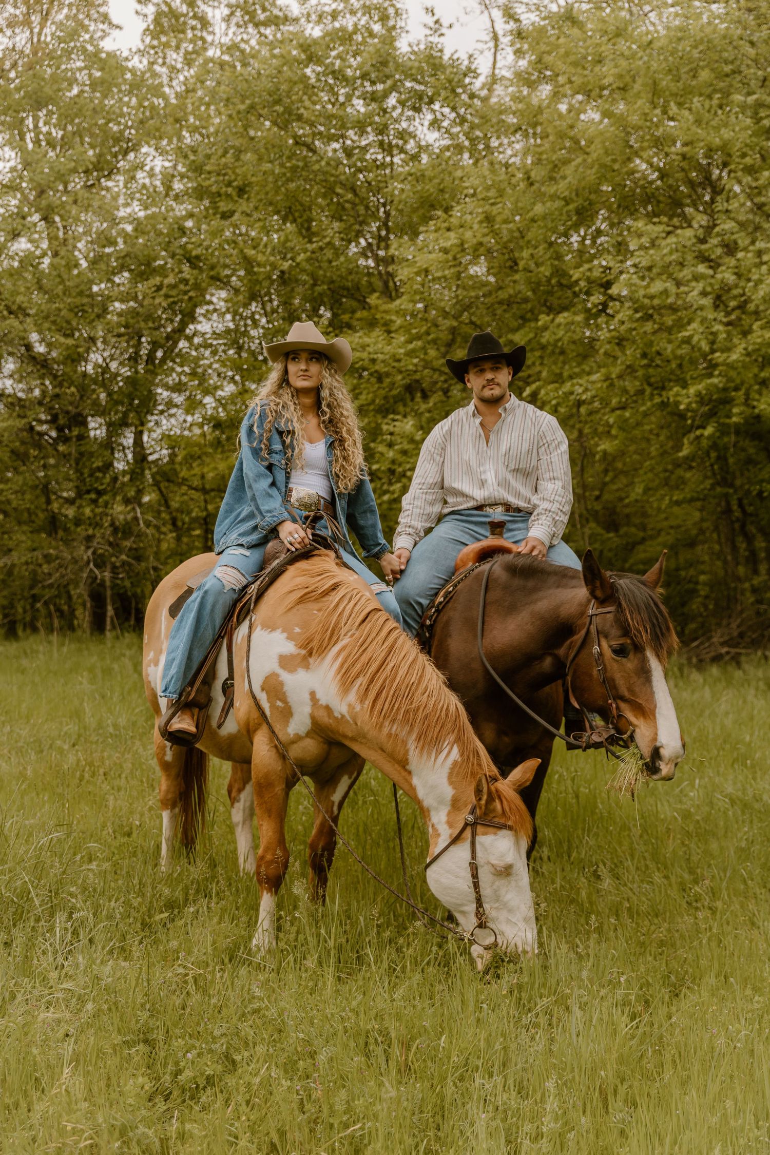 Two people ride horses through a green meadow wearing western attire and cowboy hats.