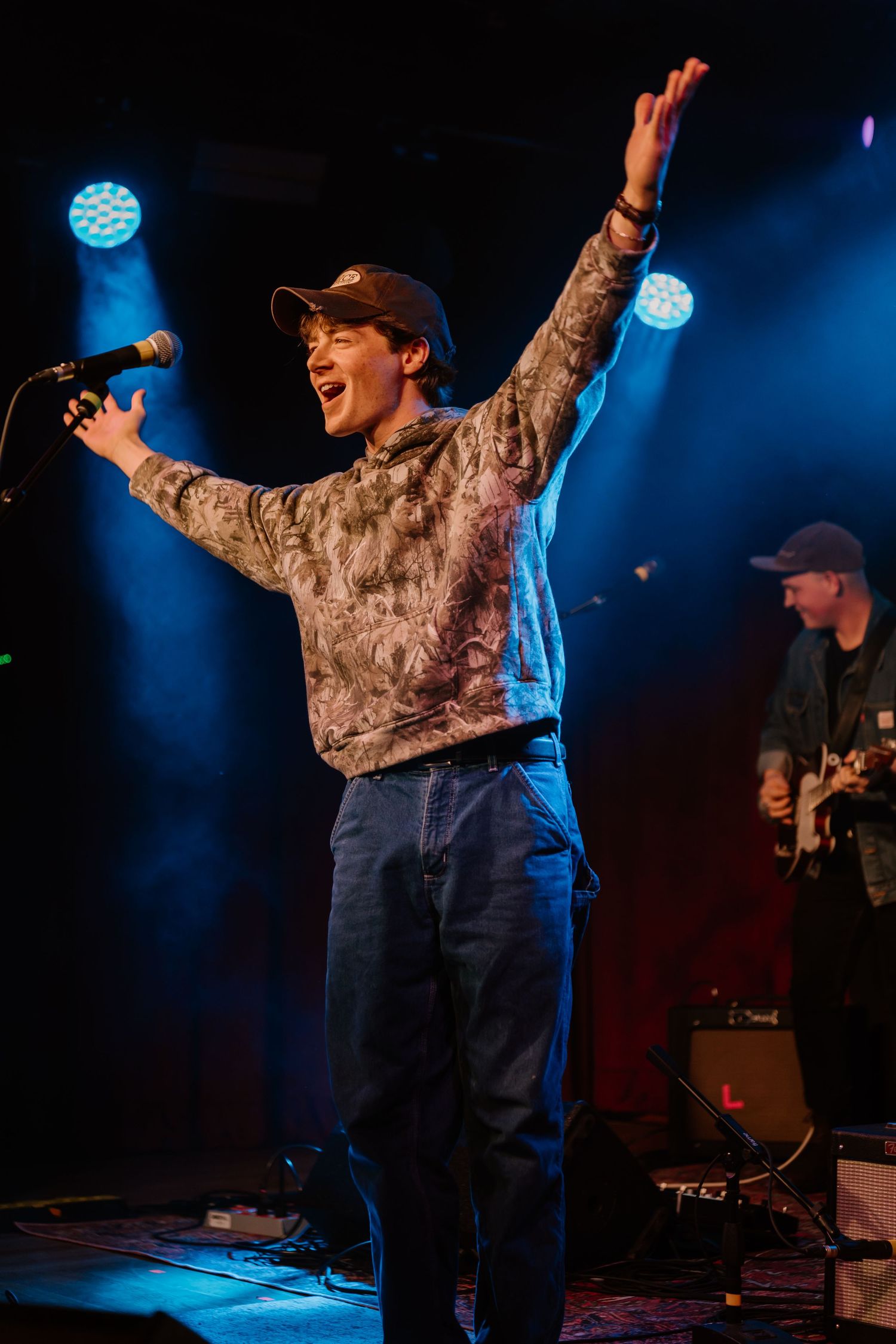 Performer in camo sweatshirt sings passionately on stage with blue lighting.