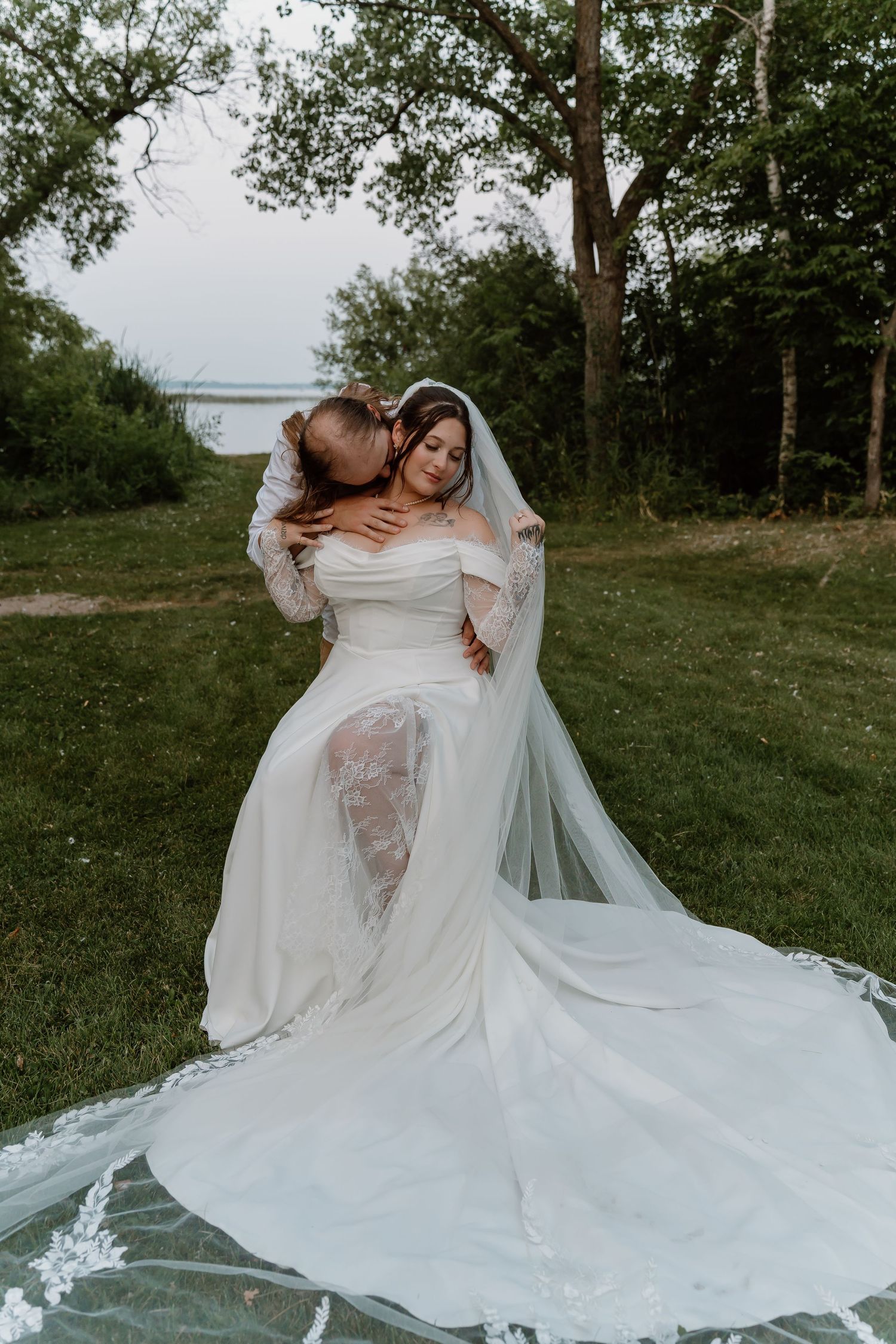 Two brides embrace outdoors in white wedding gowns, one wearing a long veil, surrounded by lush greenery near a lake.