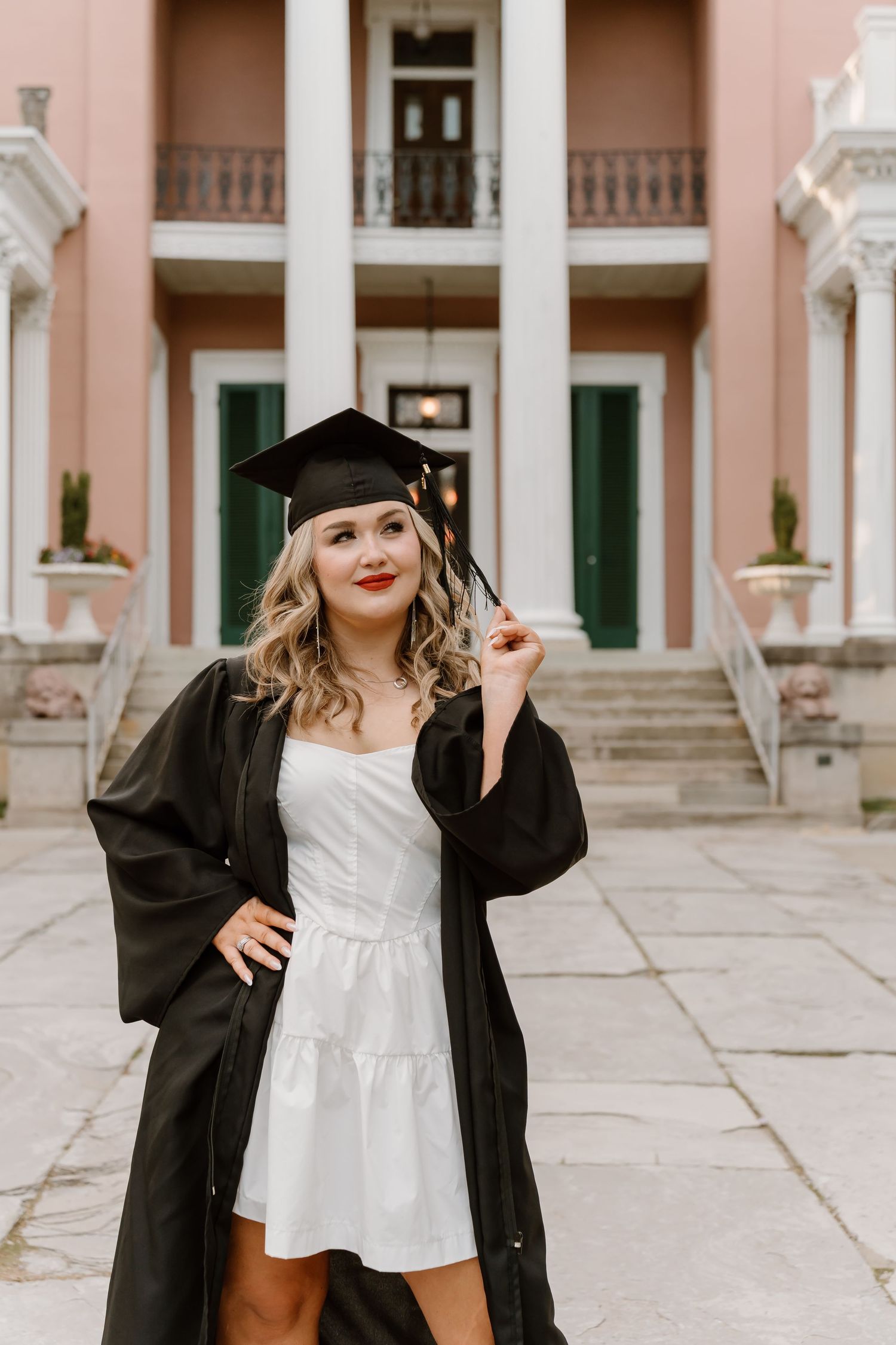 Young woman in graduation cap and gown over white dress poses proudly in front of a pink columned building.