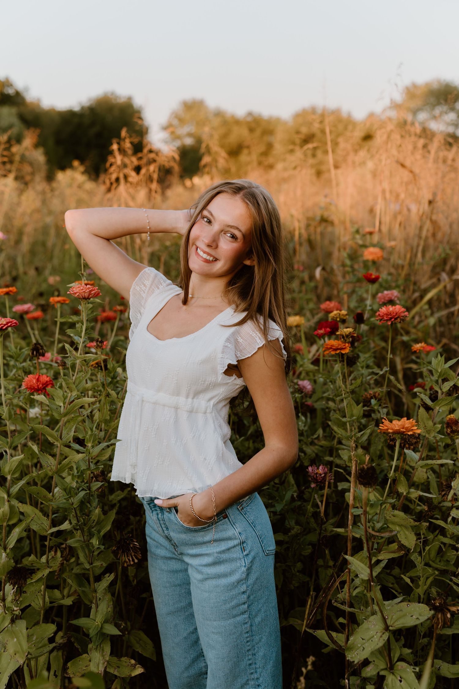 Portrait of woman in white top and jeans standing among red zinnias at golden hour.