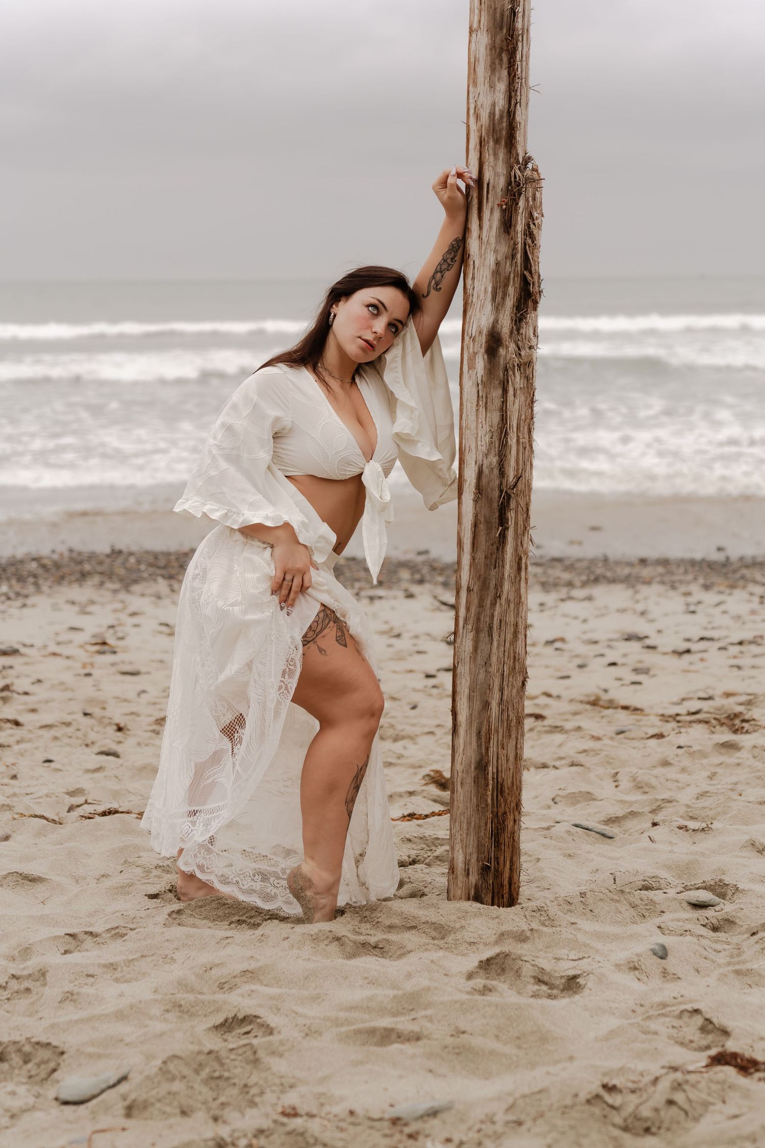 Woman in white lace crop top and skirt posing against a wooden pole on a sandy beach with ocean waves.