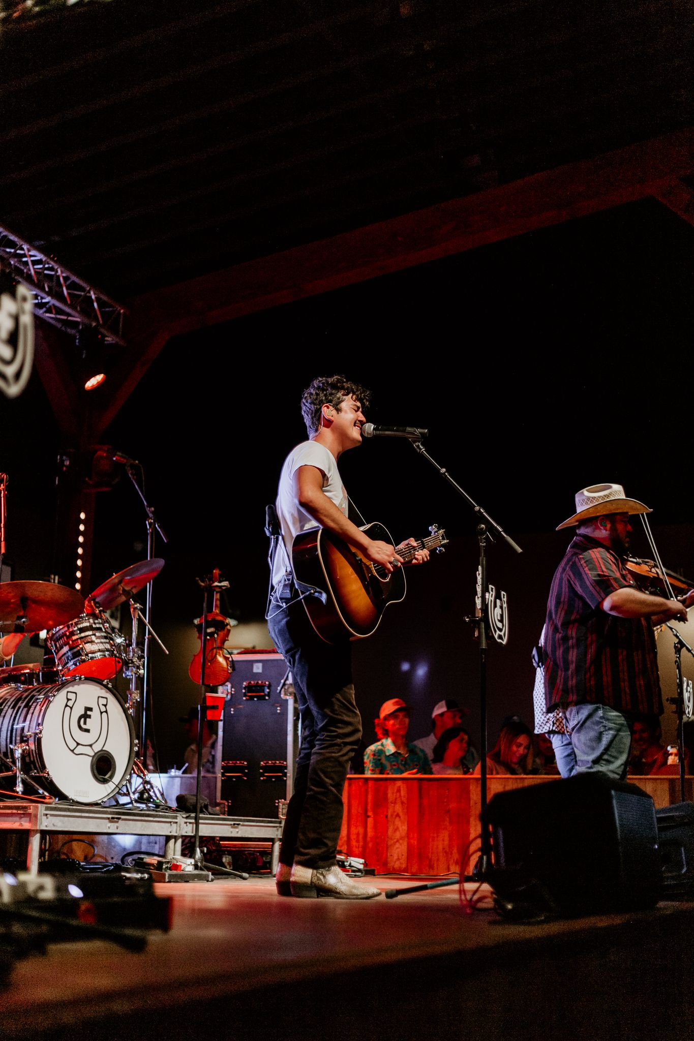 Musicians perform on a dimly lit stage with acoustic guitars and drums during a live concert performance.