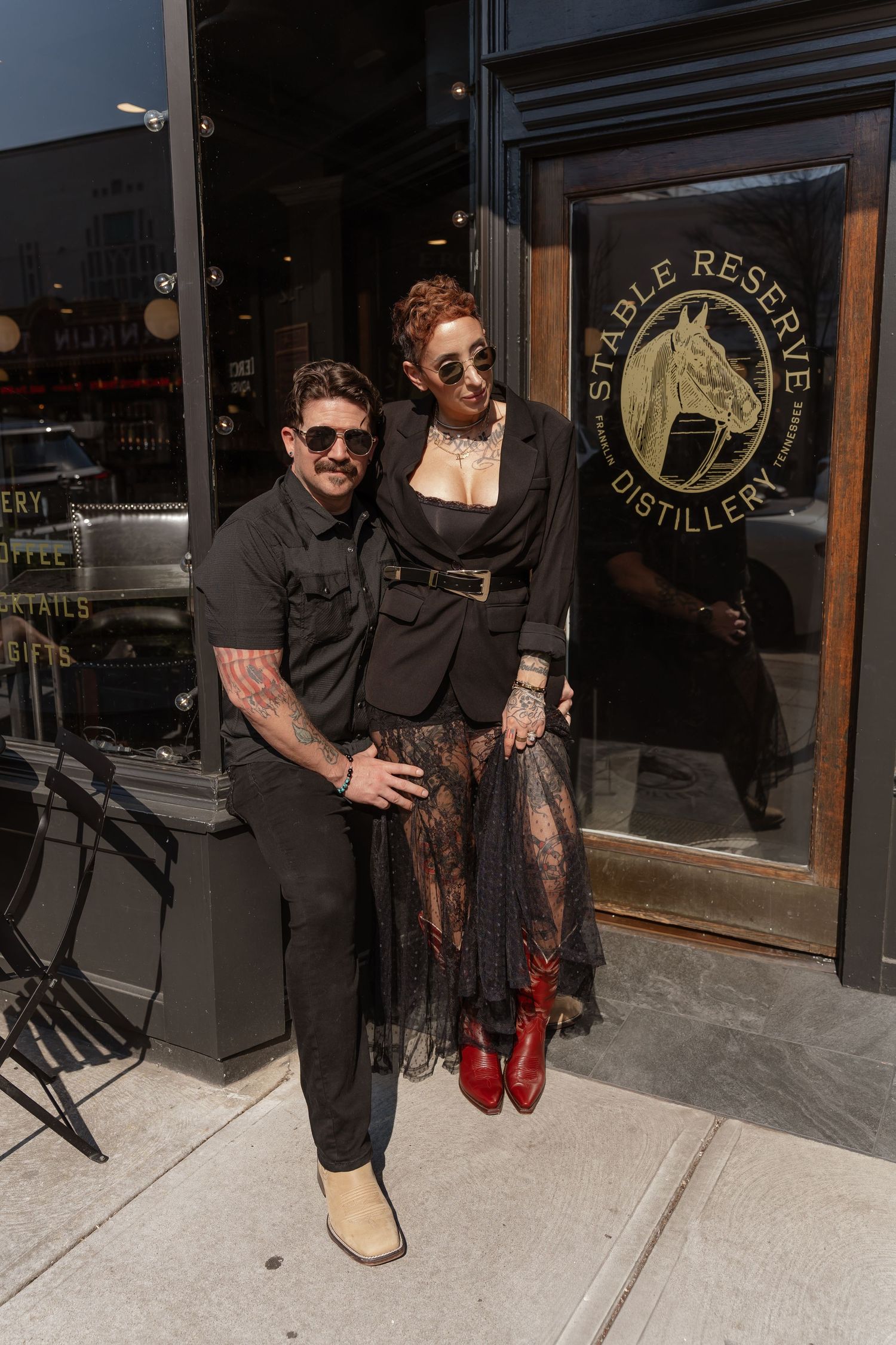 Two people in dark outfits pose together outside Stable Reserve Distillery with a horse logo visible on the door.