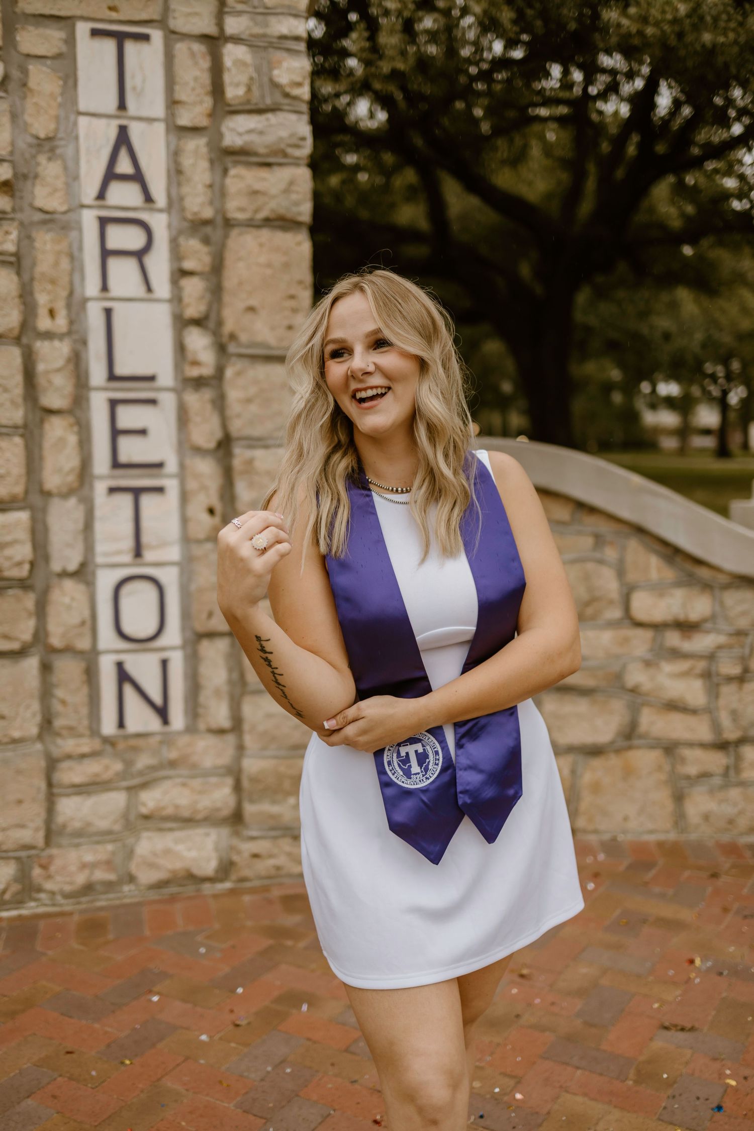 A graduate in a white dress and purple sash poses by a stone Tarleton sign.