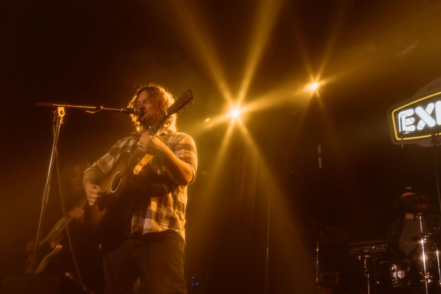 Male guitarist performs on stage under golden spotlights at the Exchange venue, playing acoustic guitar at microphone.