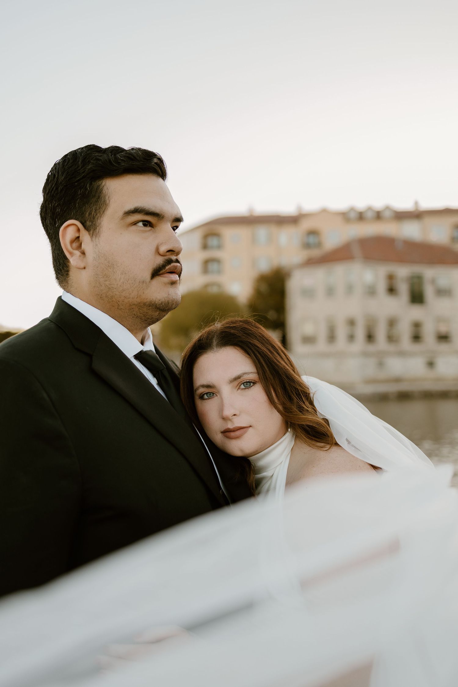 Romantic wedding couple poses together against a blurred architectural backdrop in warm lighting.