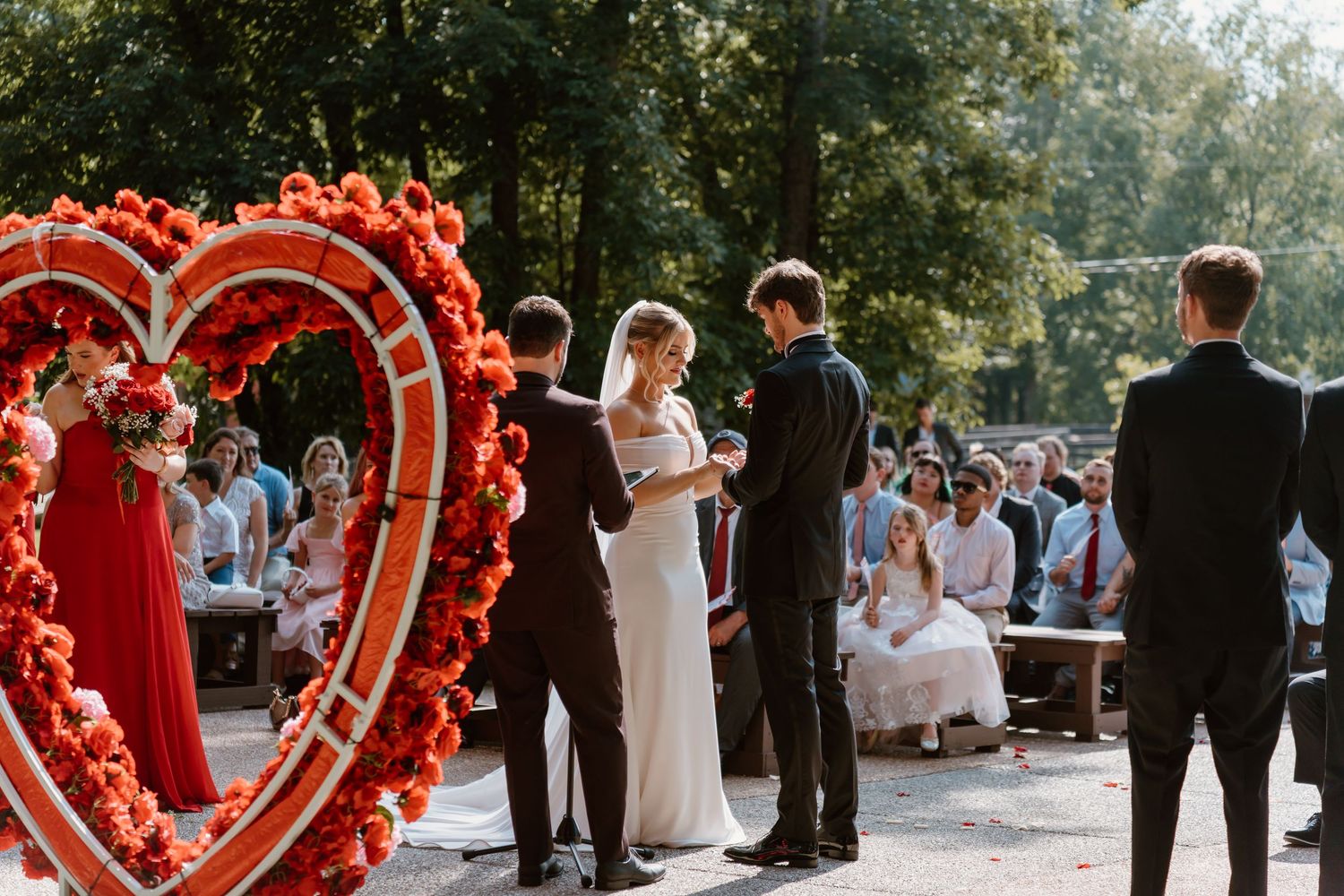 Outdoor wedding ceremony setup with a large red heart-shaped floral arch and guests gathered in a garden setting.