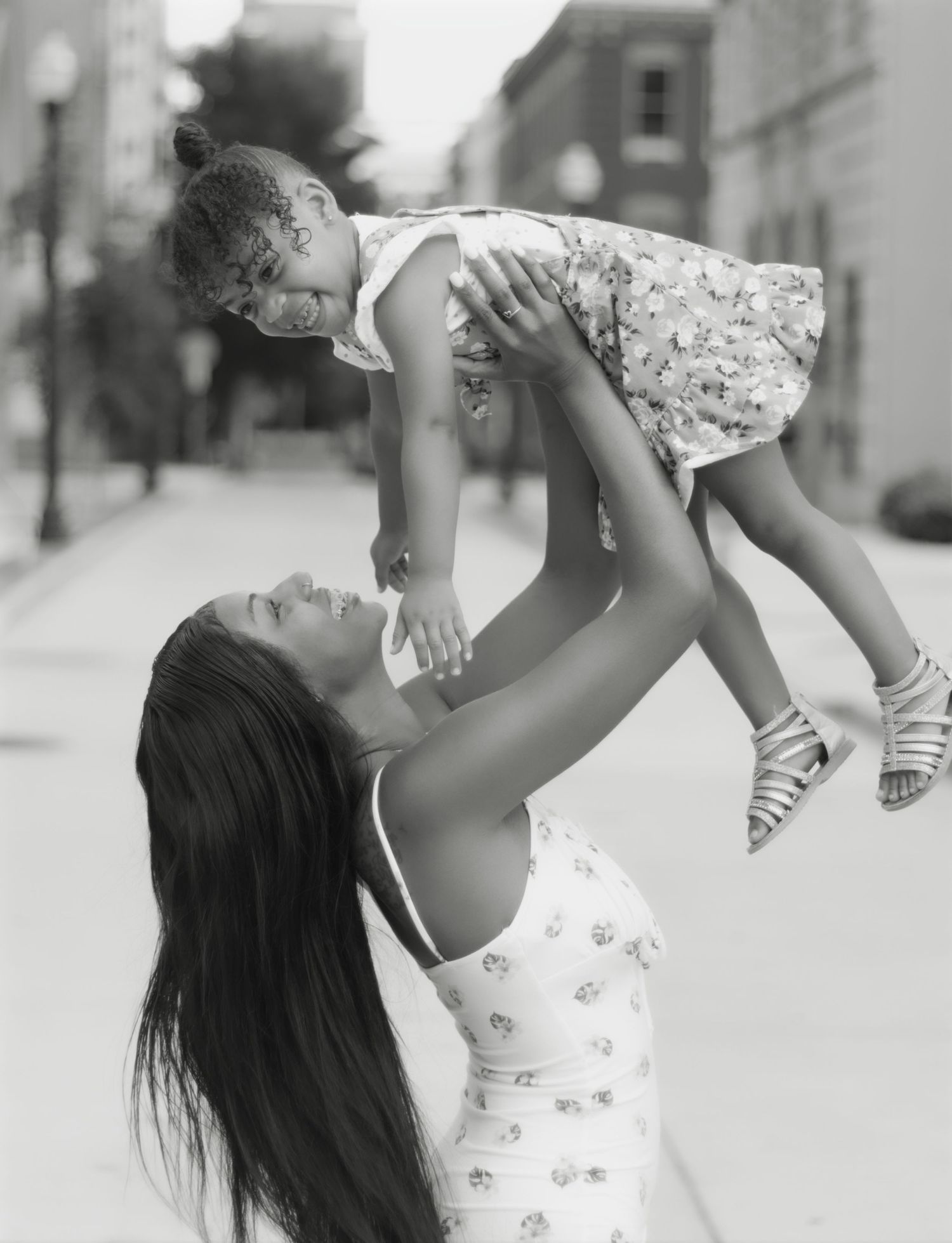 Black and white photo of parent lifting child in floral dress on city street.
