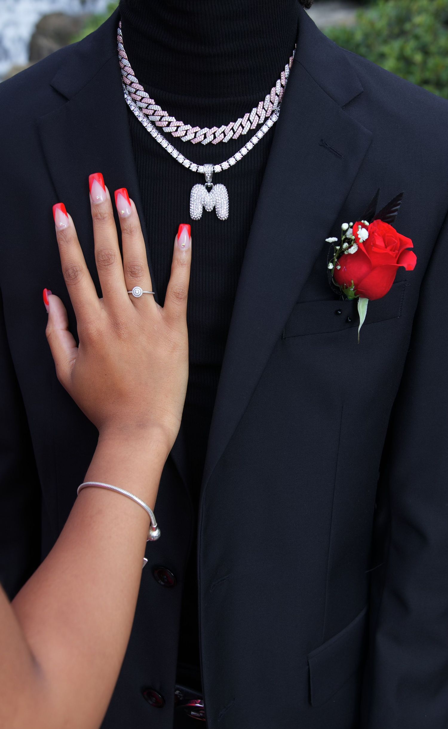 Close-up detail of a formal suit with a red rose boutonniere and jewelry-adorned hand resting on lapel.