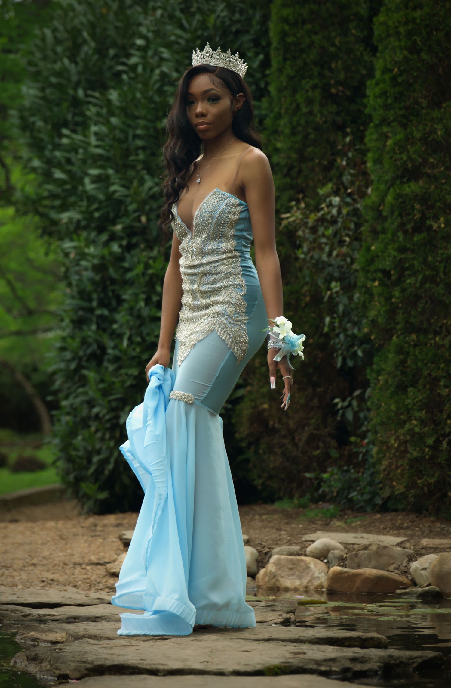 Woman in light blue formal gown and crown posing in garden setting.