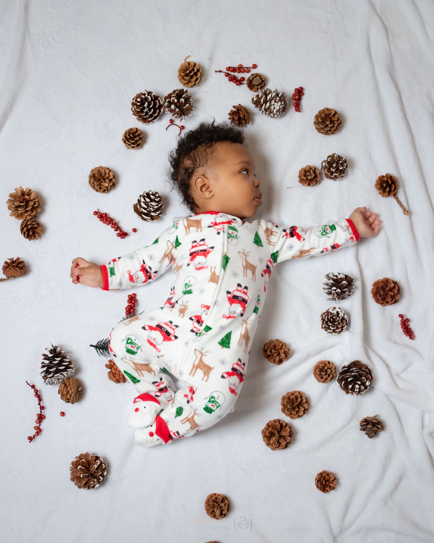 Baby wearing Christmas pajamas lies surrounded by pinecones and red berries on white bedding.