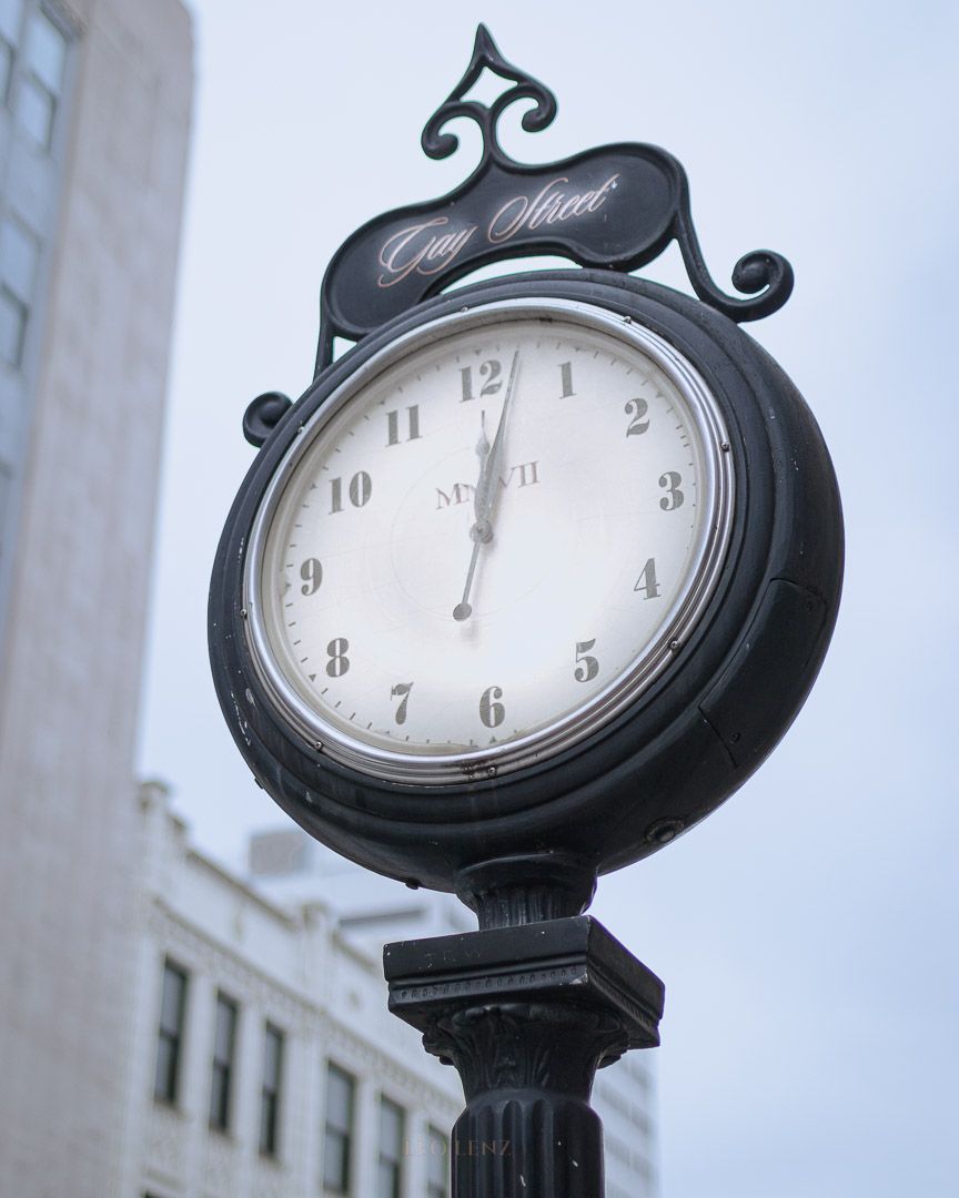 Vintage-style street clock mounted on decorative black post against urban backdrop.
