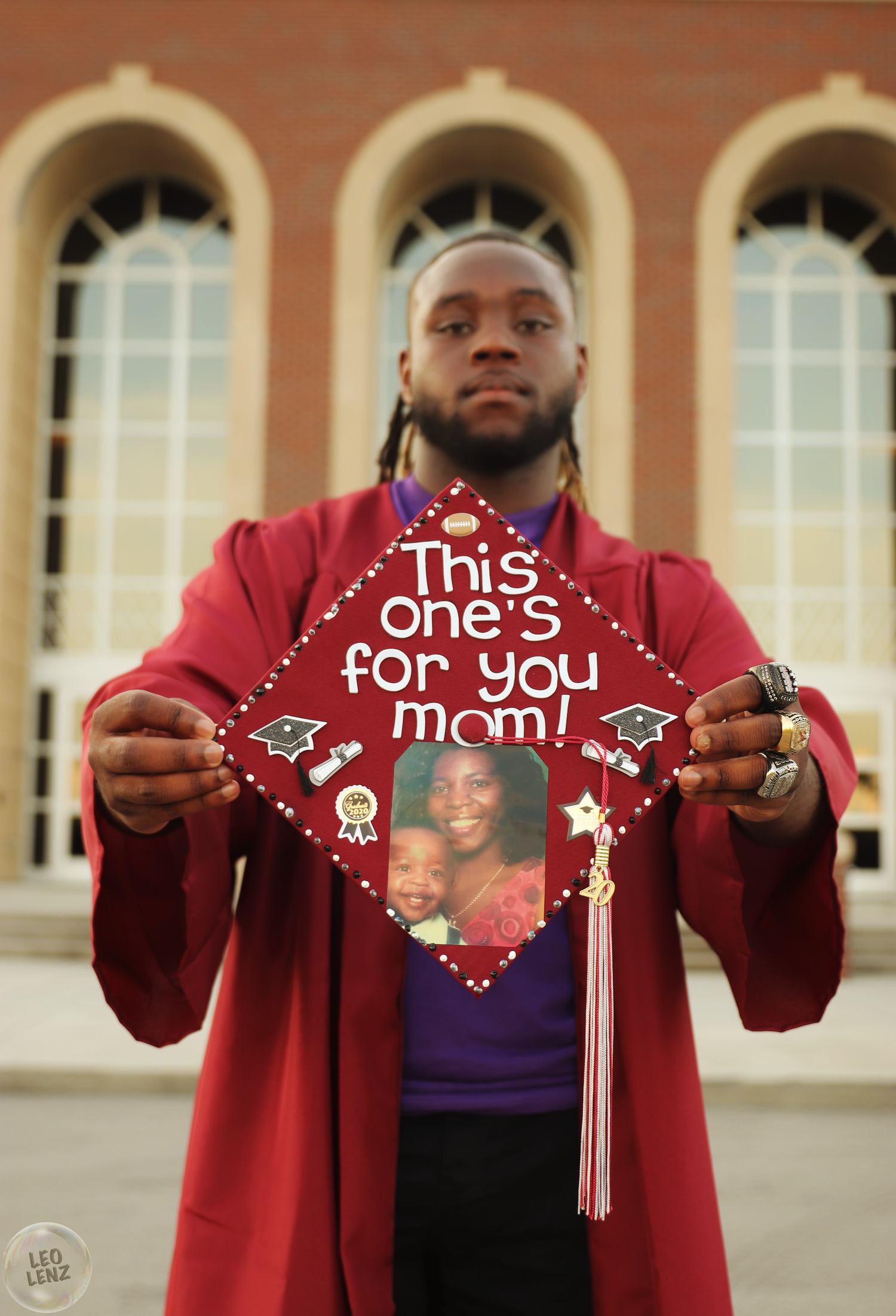 Graduate in red robe displaying decorated graduation caps with sentimental messages.