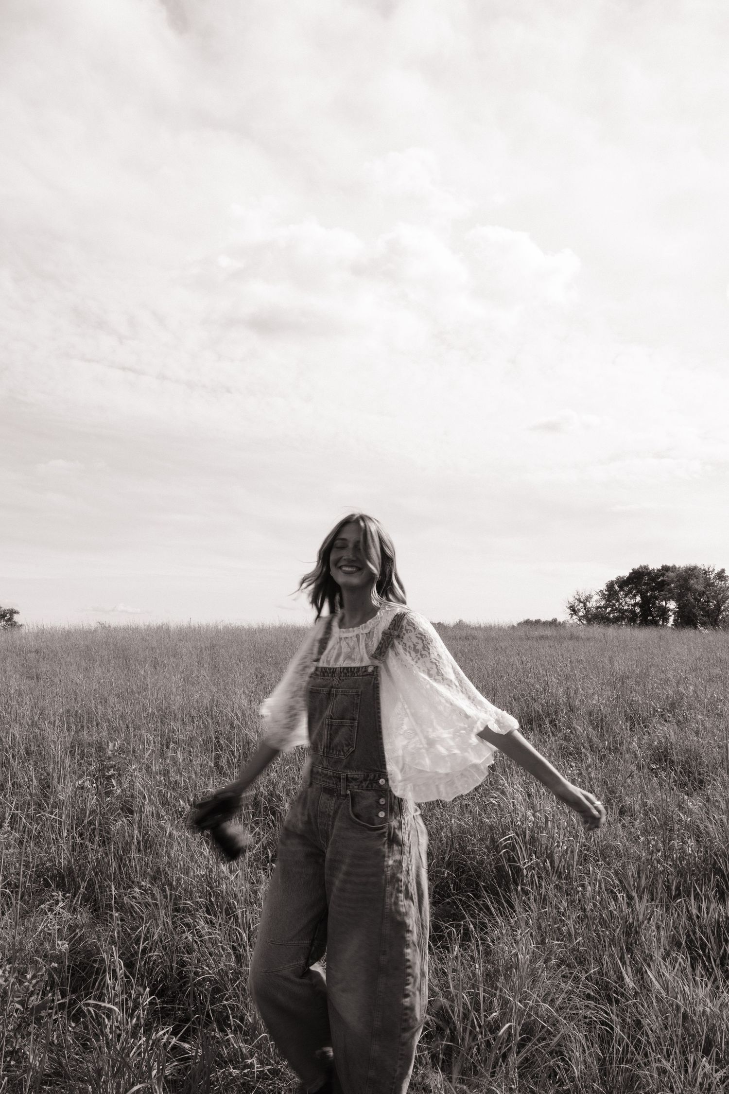 Black and white photo of a joyful woman in overalls and lace top dancing in a field under cloudy skies.
