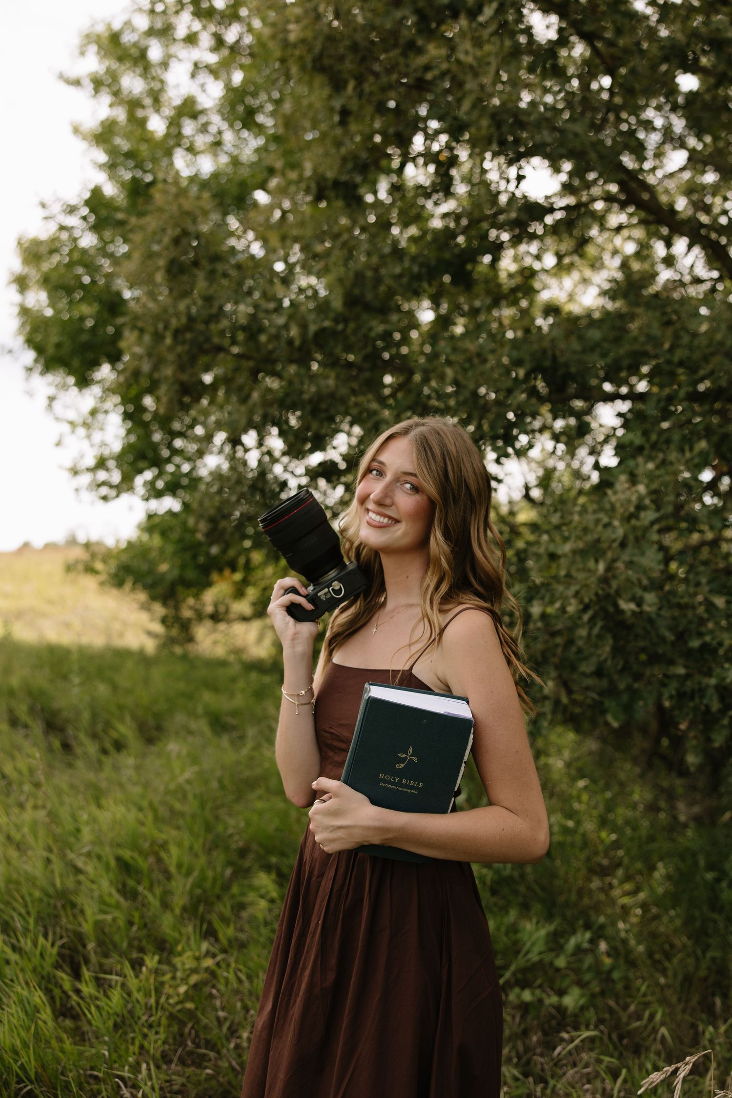 Young woman in brown dress holding camera and book smiling outdoors among green trees and grass.