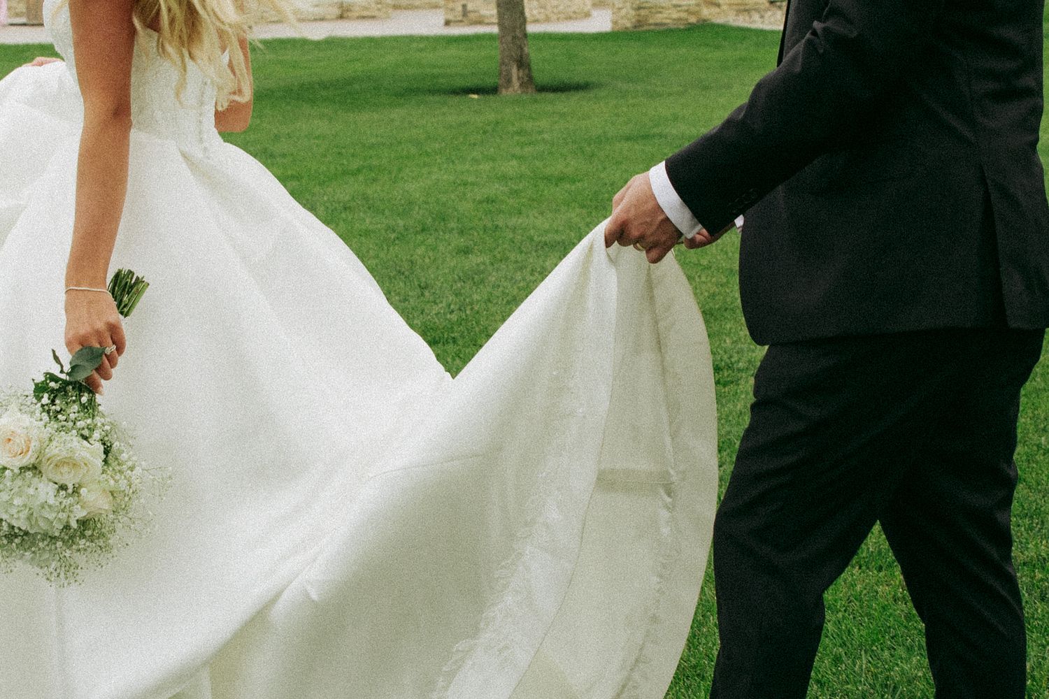 Bride in a flowing white gown holding a bouquet walks across a green lawn beside a groom in a dark suit holding her train.