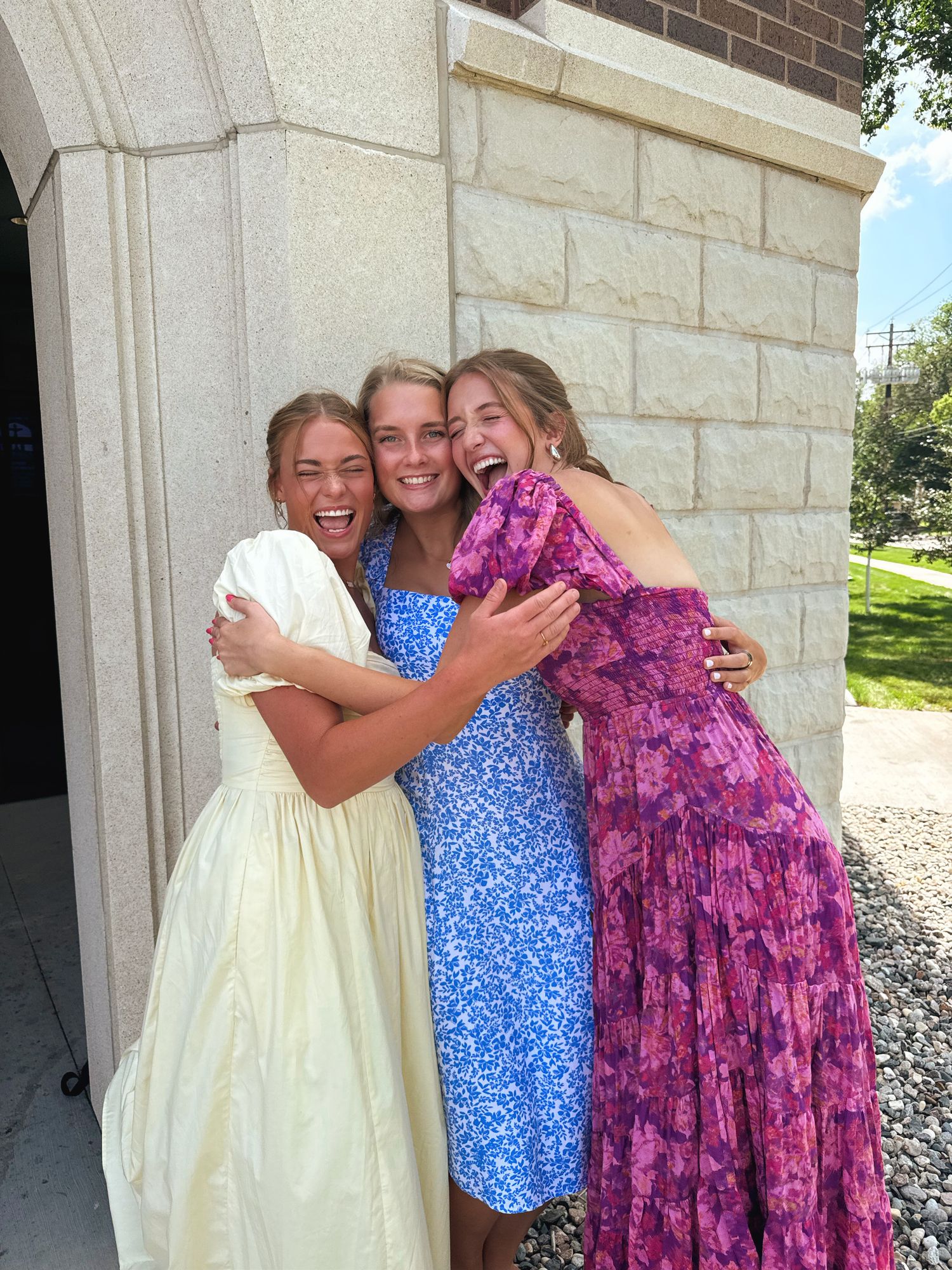 Three young women in colorful dresses laughing and hugging outside a stone building.