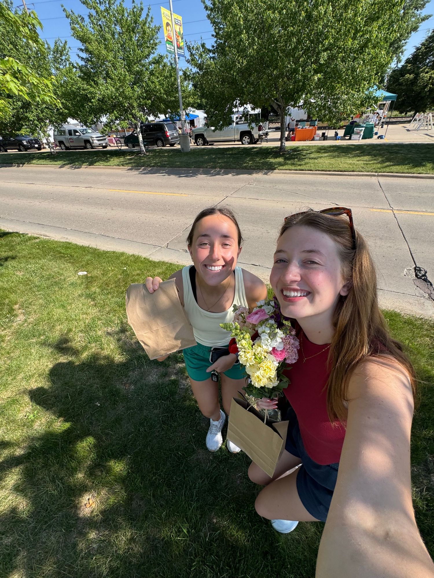Two smiling young women take a selfie outdoors on a sunny day, one holding a bouquet of flowers near a road.