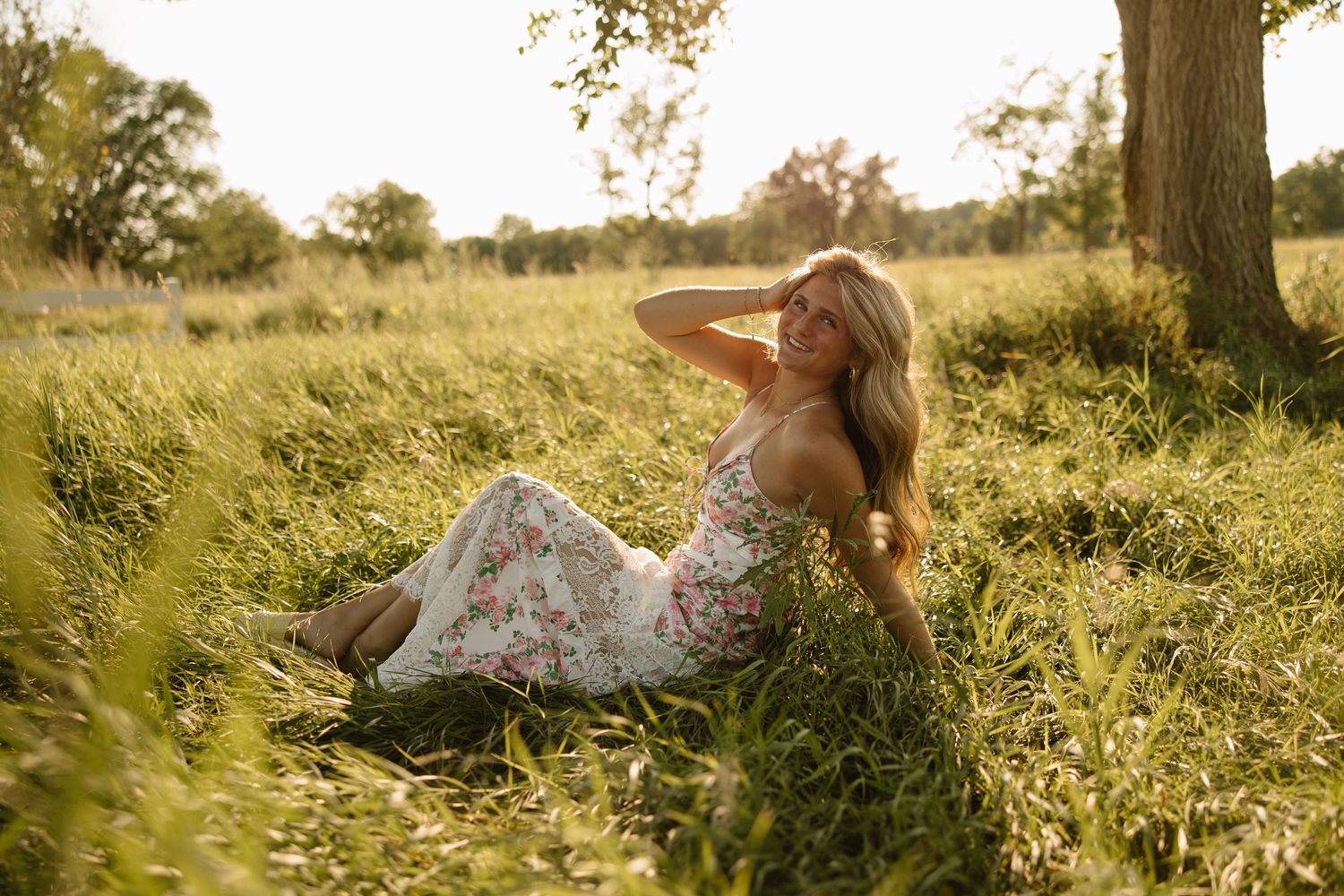 A young woman in a floral dress sits in a sunlit grassy field, resting her hand on her head near a tree.