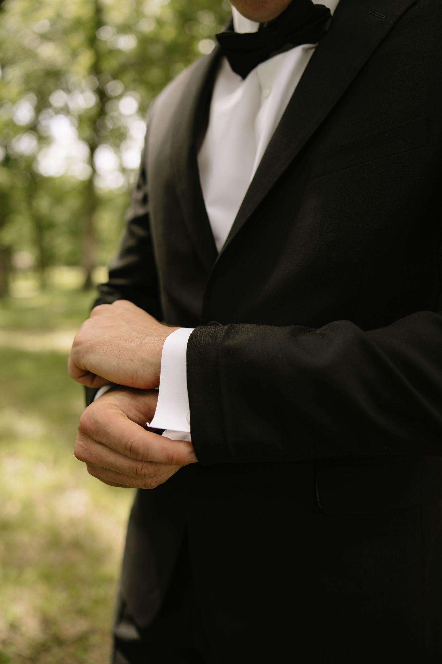 A groom in a black tuxedo adjusts his cufflinks outdoors with green trees in the background.