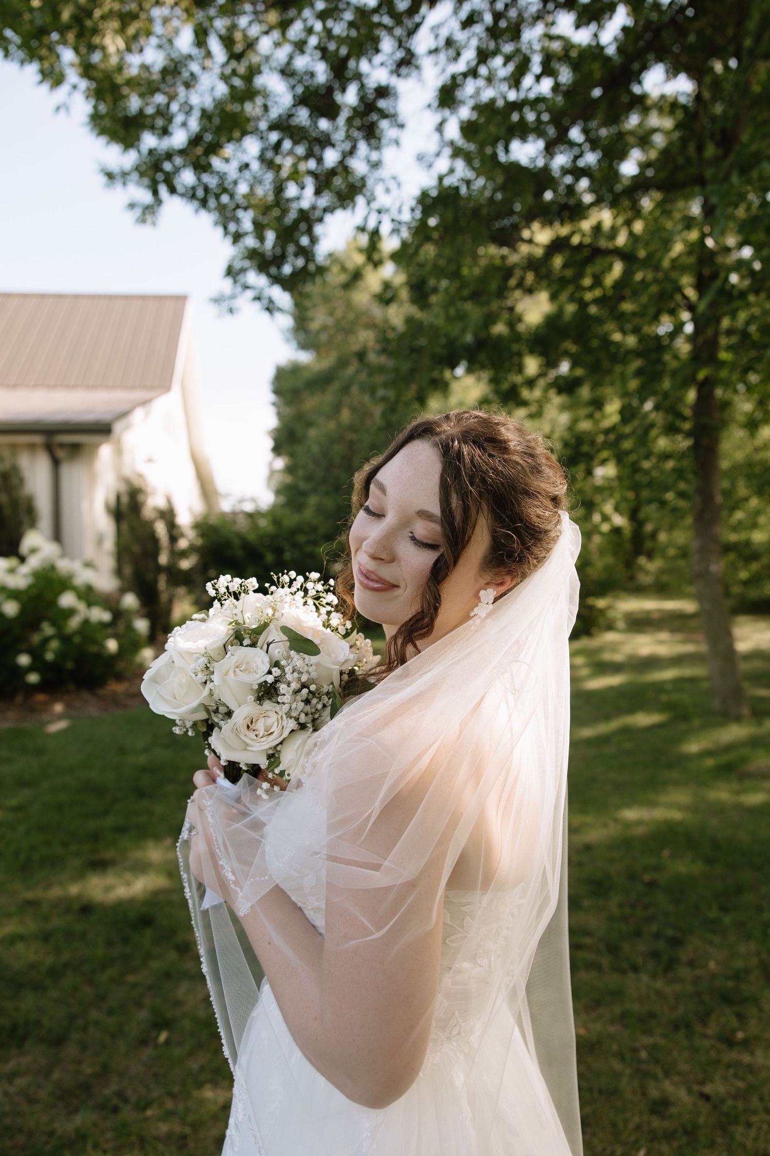 Bride in white gown and veil holds bouquet of white roses outdoors, smiling serenely in a lush green garden setting.