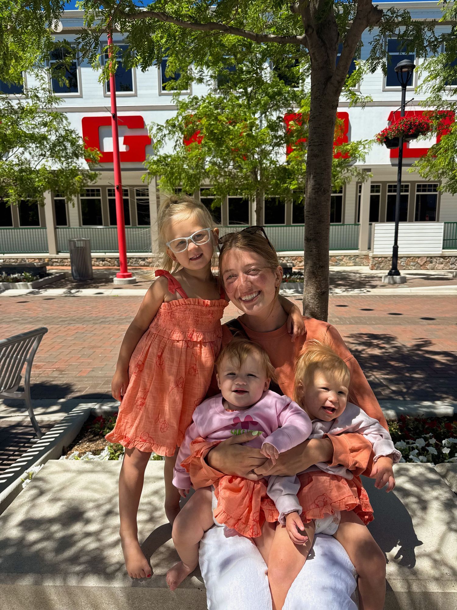 Woman in orange dress sits on bench holding two babies while older girl in matching outfit poses beside her outdoors.