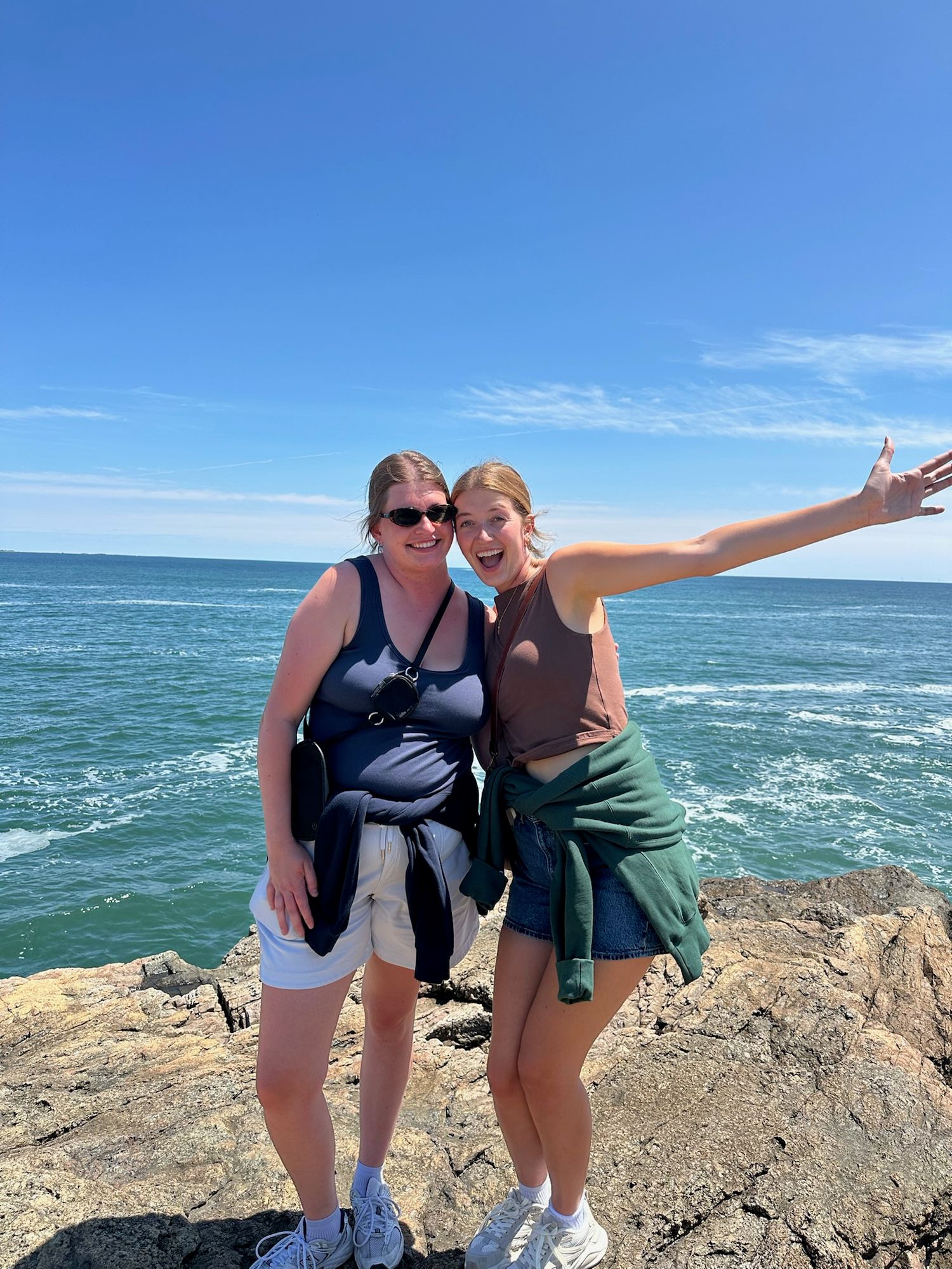 Two women posing on rocky coastal cliffs with ocean waves and clear blue sky in the background on a sunny day.