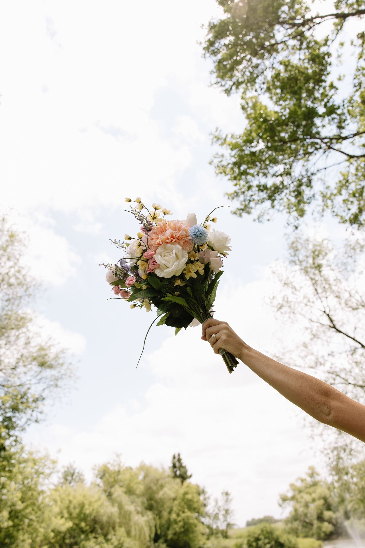 Hand holding a colorful bridal bouquet with white, pink, and blue flowers raised toward a cloudy sky.