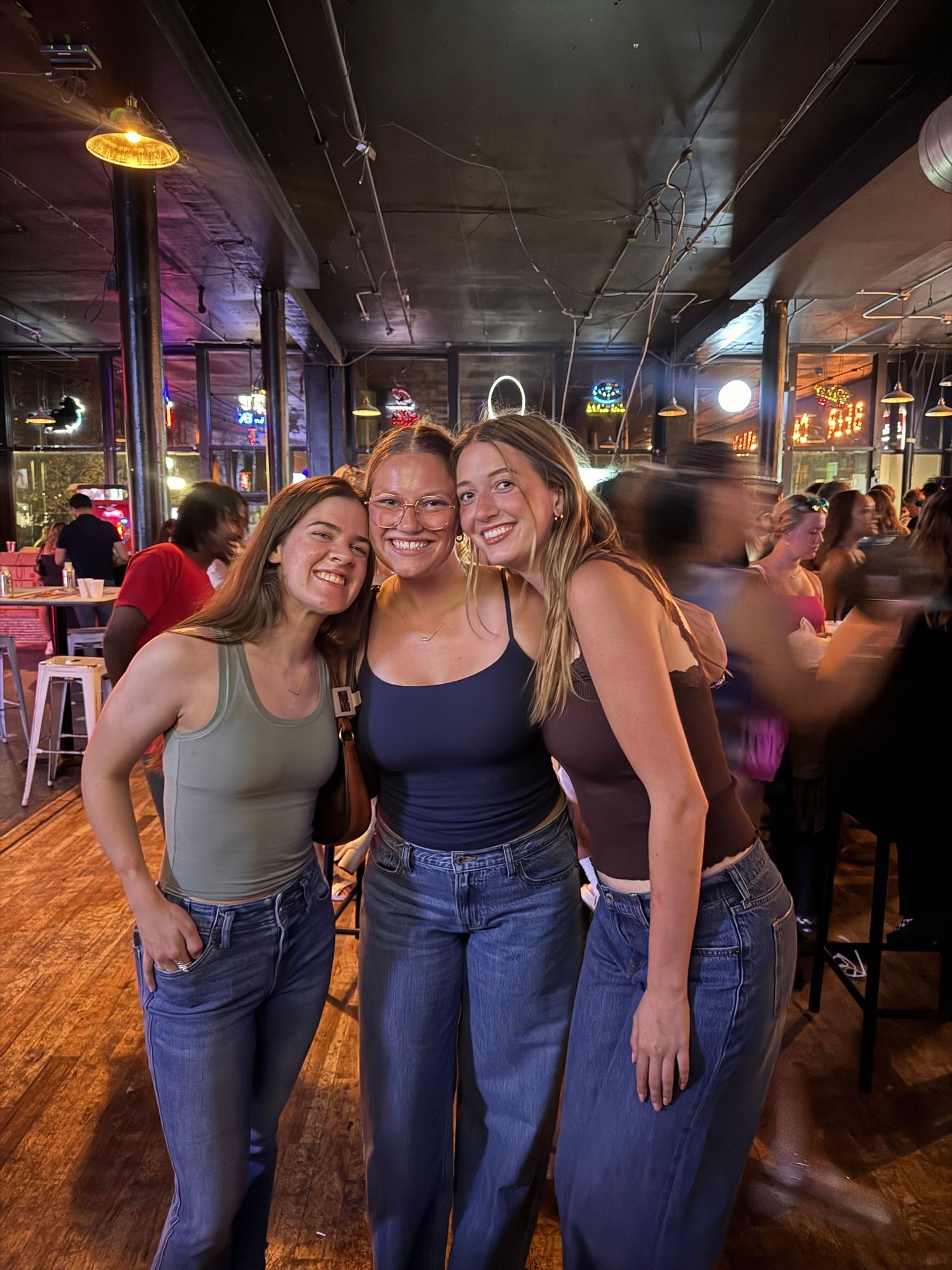 Three smiling young women pose together in a lively bar with warm lighting and neon signs.
