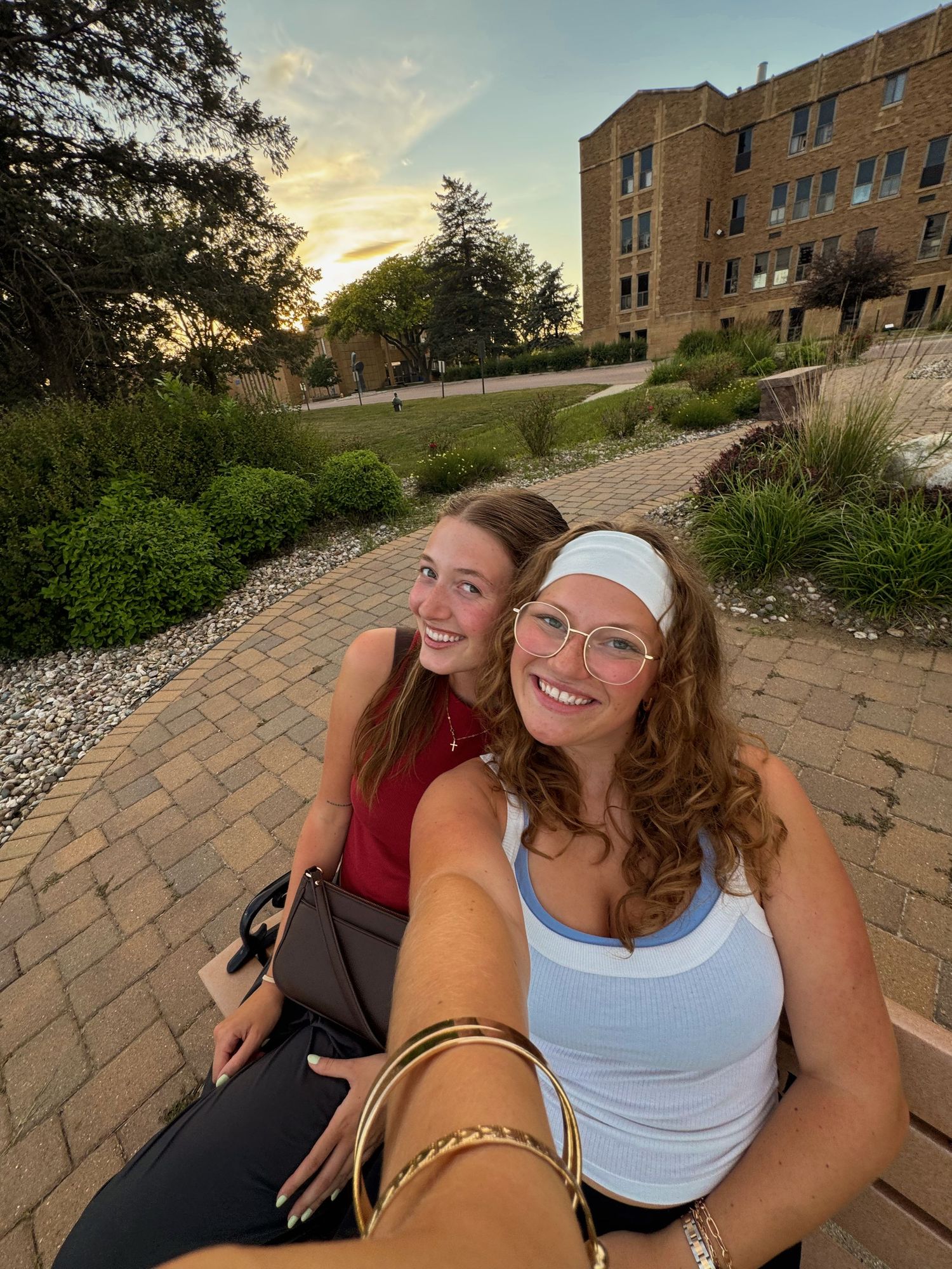 Two smiling young women take a selfie on a brick path at sunset near a college campus building.