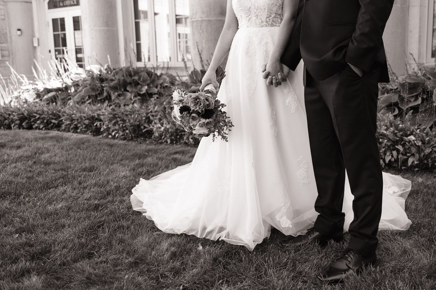 Black and white photo of a bride in a flowing tulle gown holding a bouquet beside a groom in a dark suit on a manicured lawn.