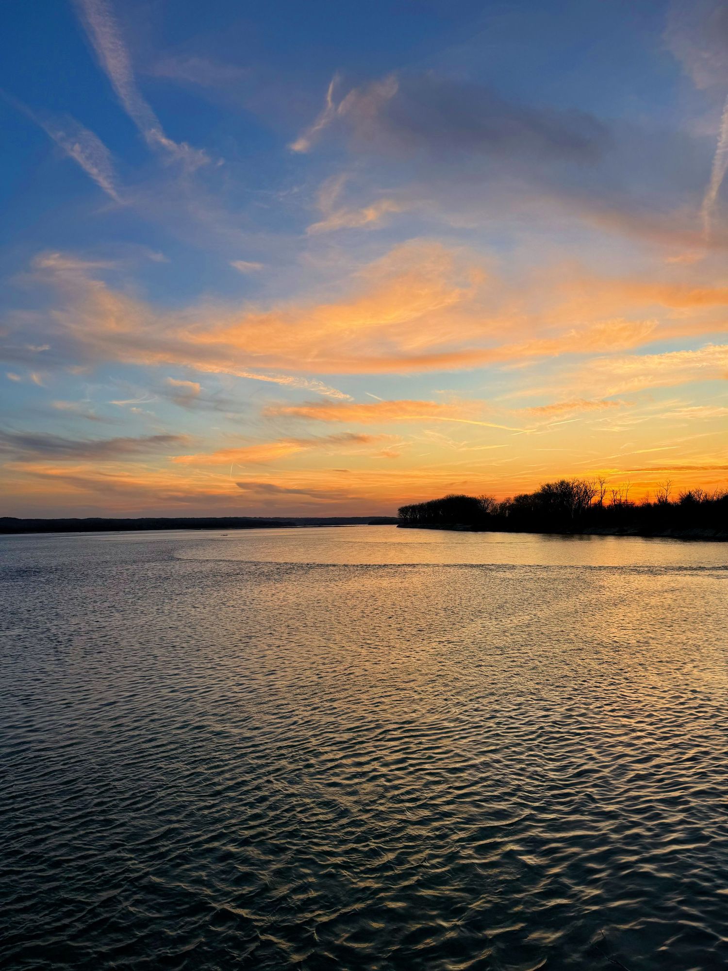 Vibrant sunset over a calm river with orange and blue skies reflecting on the water near a silhouetted tree line.