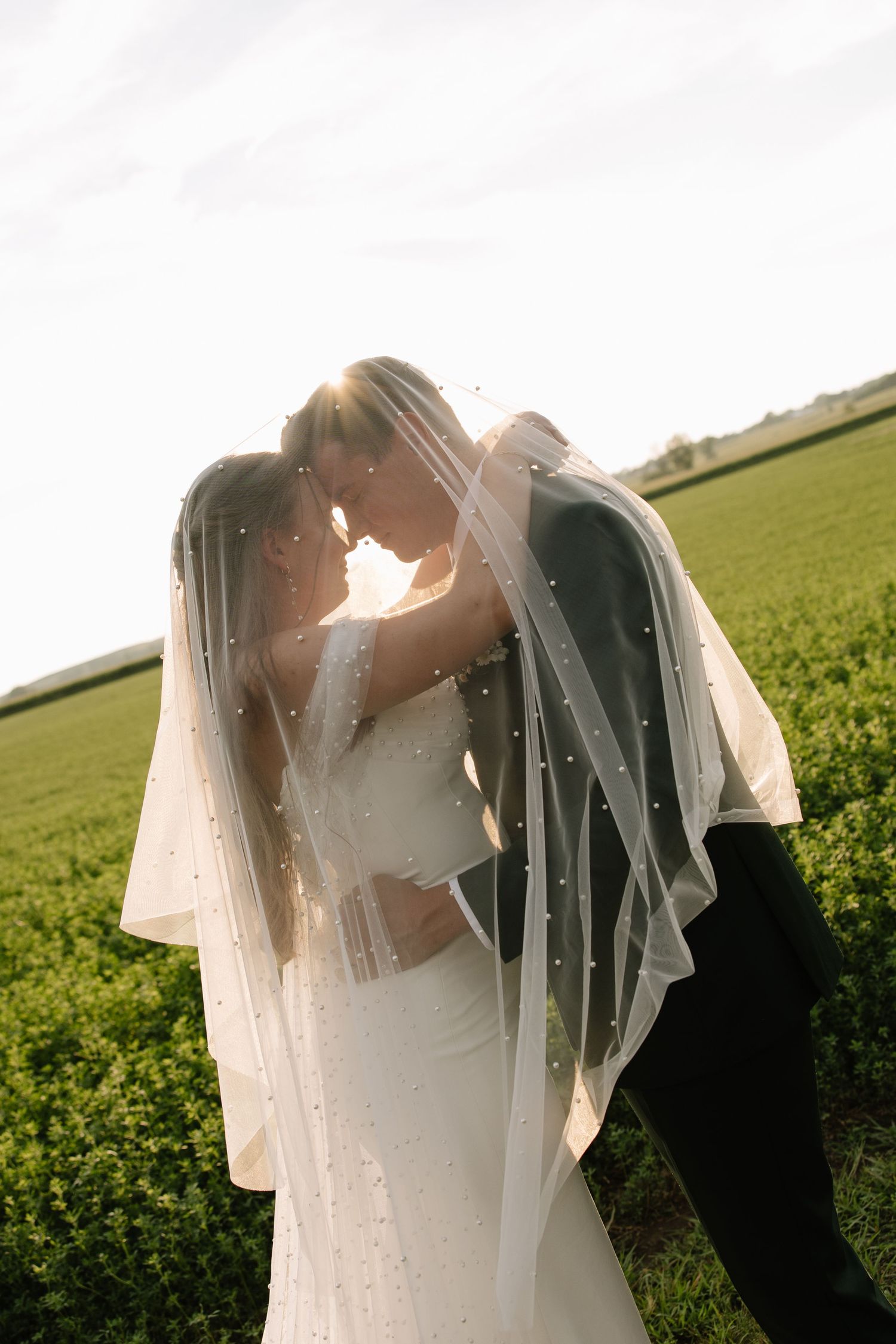 Bride and groom nearly kissing under pearl-adorned veil in sunlit green Nebraska field.