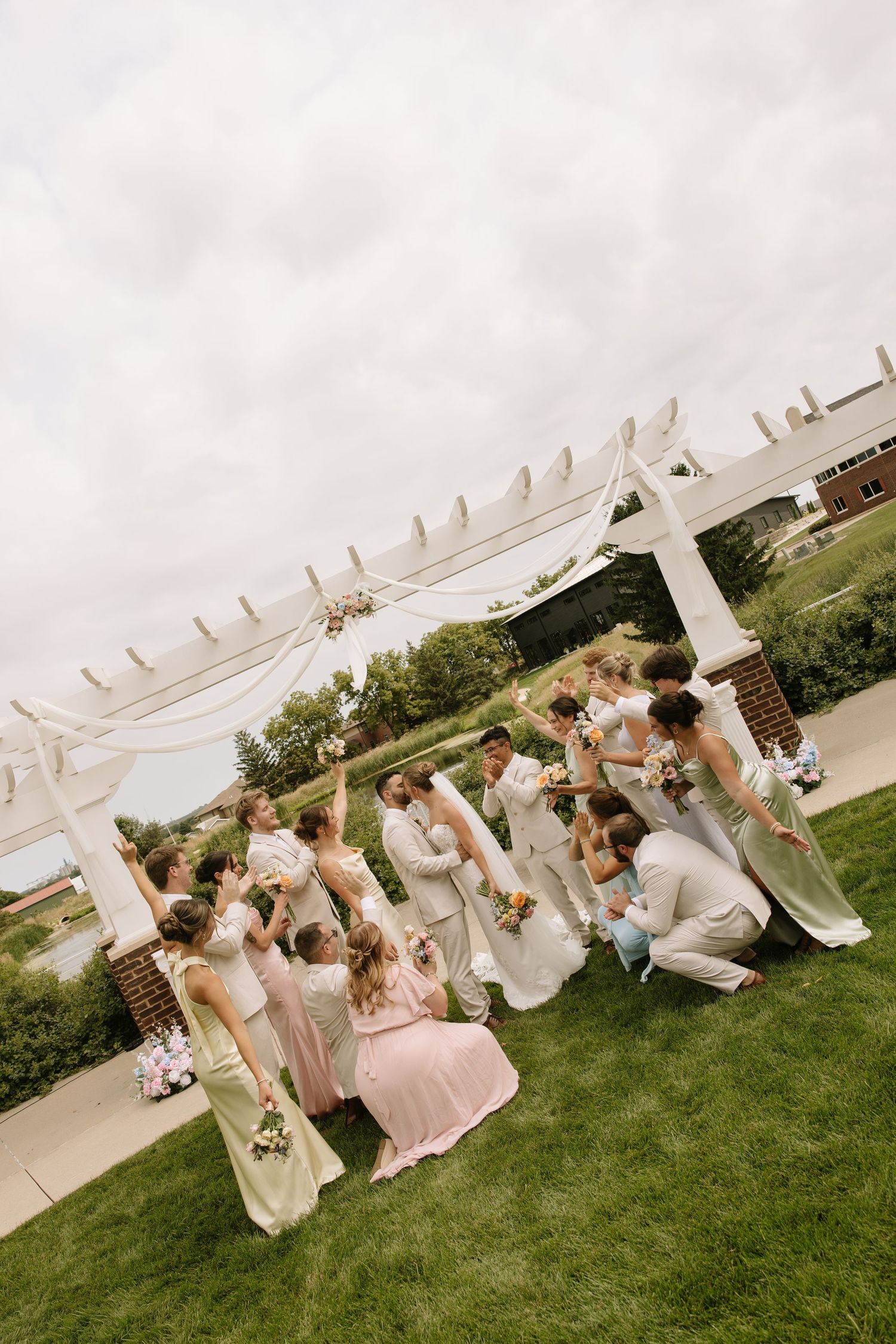 Wedding party poses under white pergola on green lawn, bridesmaids in sage and pink gowns with groomsmen in gray suits.