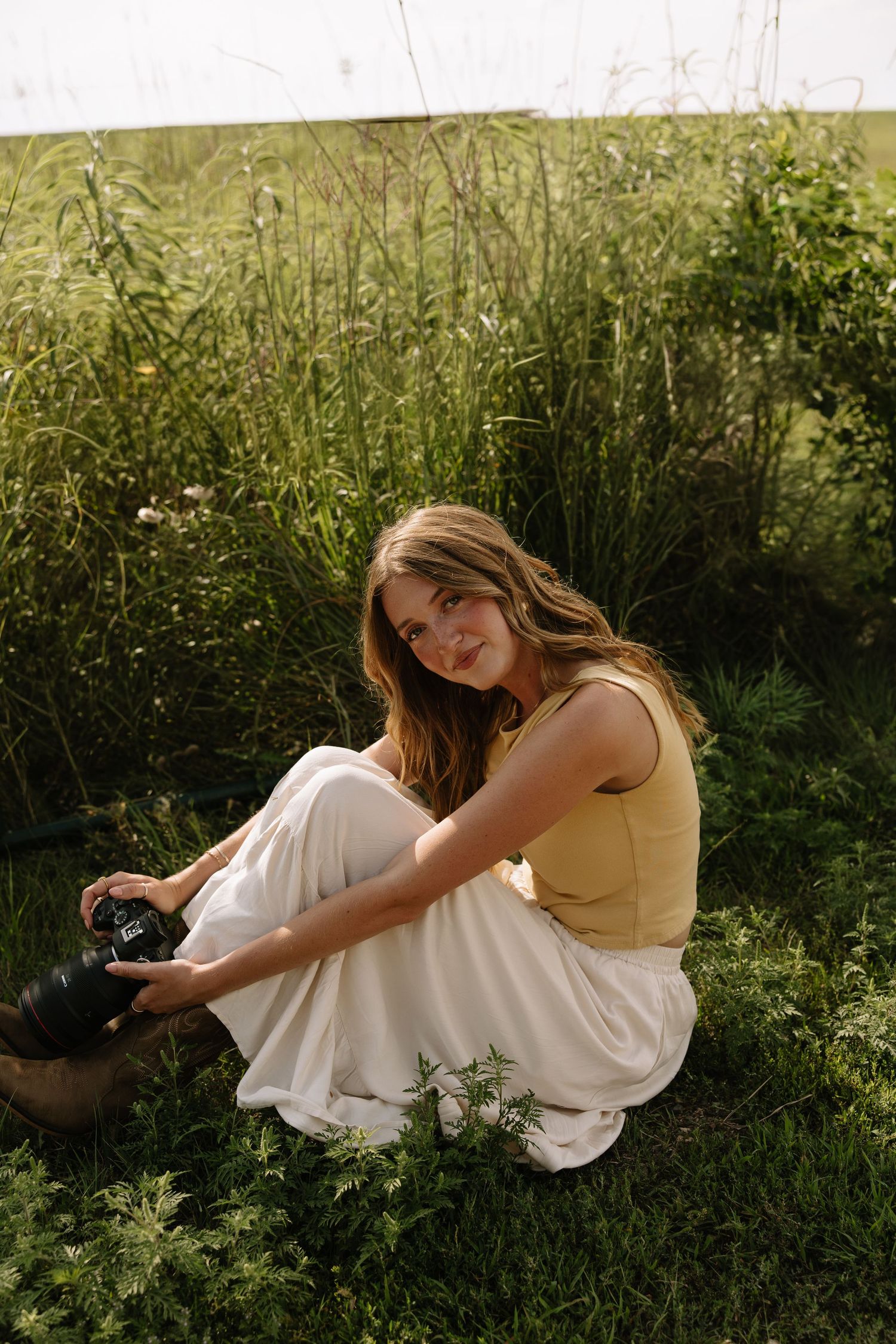 Young woman in yellow top and white skirt sitting on grass holding a camera, surrounded by lush green meadow plants.