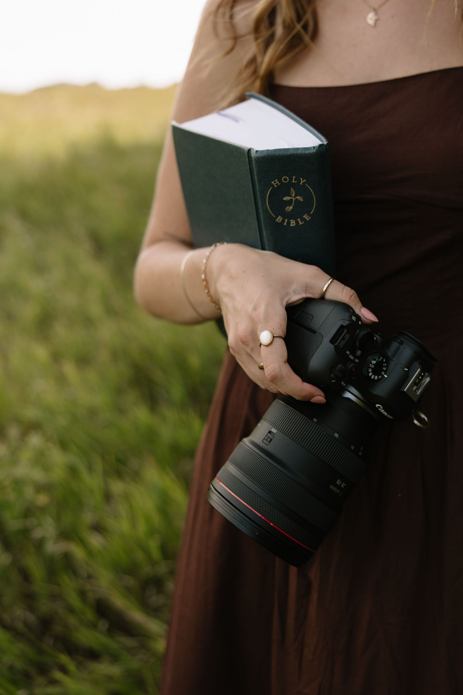 Woman in brown dress holding a Holy Bible and DSLR camera in a green field.