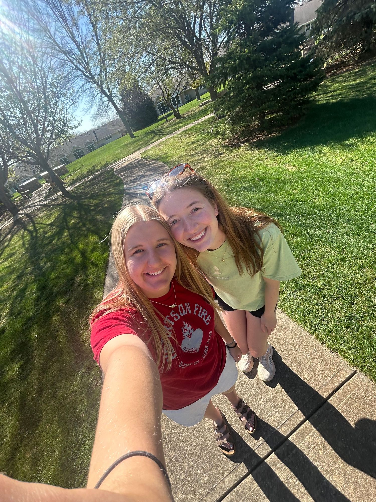 Two smiling teenage girls taking a selfie on a sunny sidewalk surrounded by green trees.