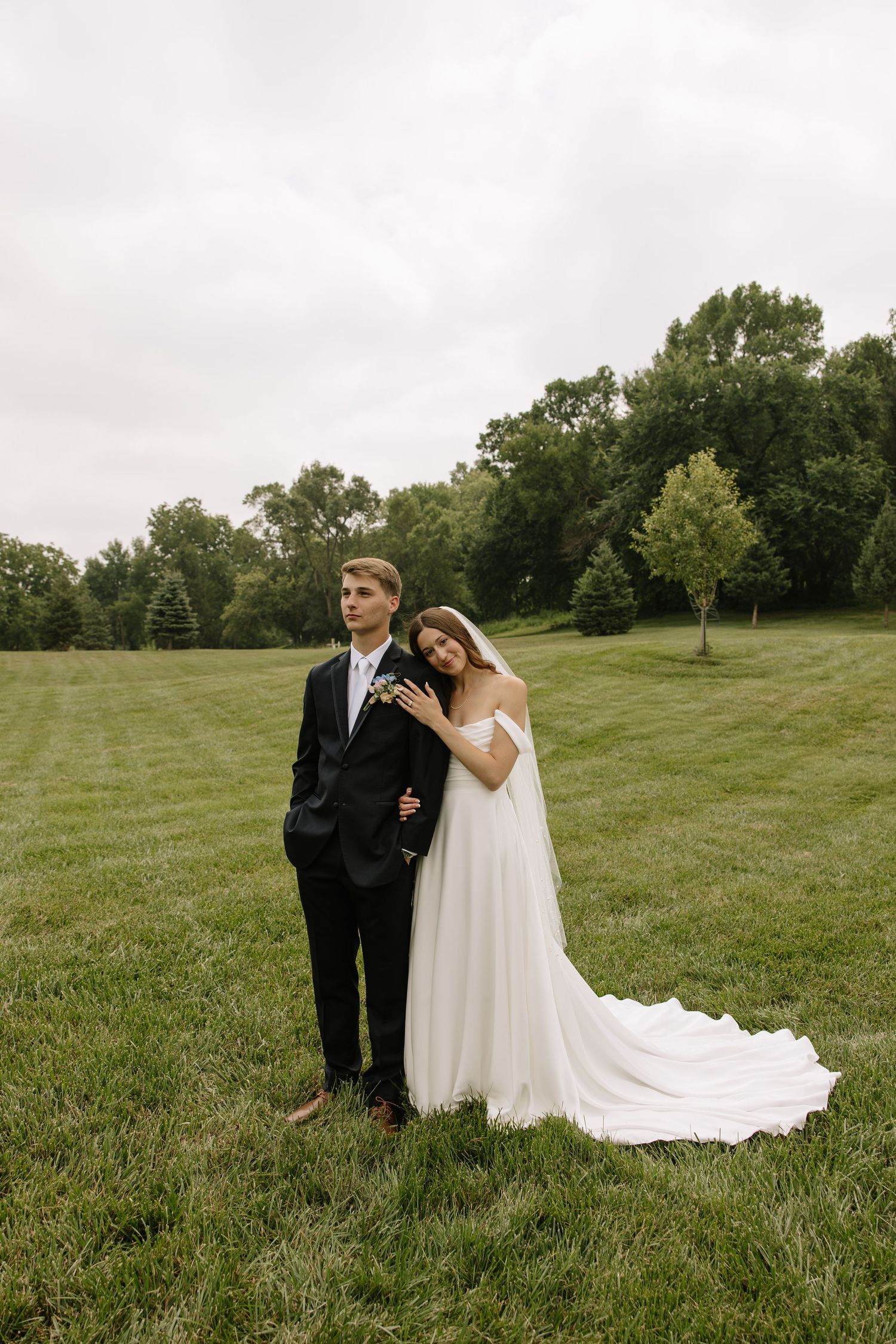 A bride in a white gown leans on a groom in a black suit, posing on a lush green lawn with trees in the background.