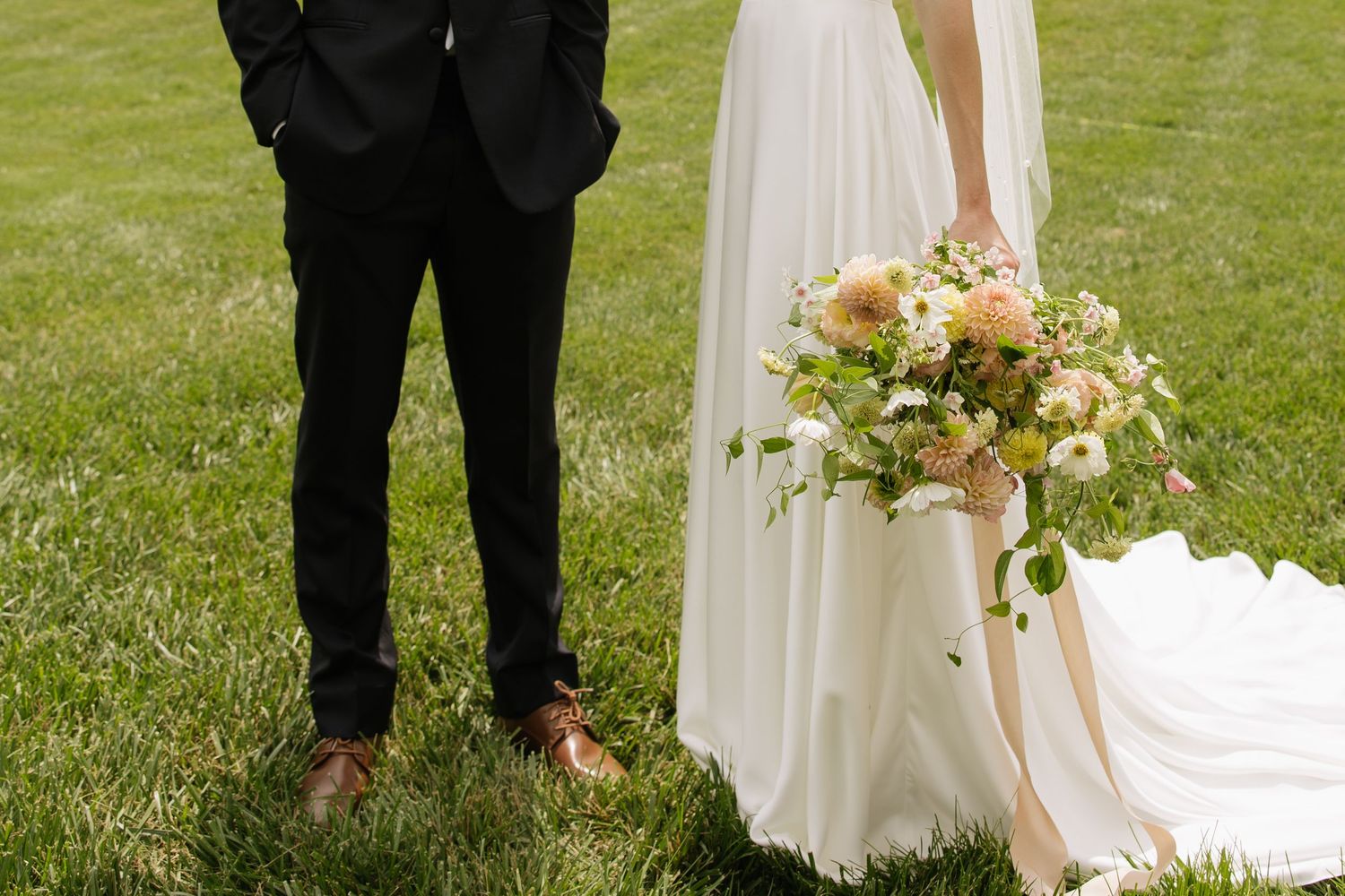 Bride in white gown holding peach floral bouquet standing beside groom in black suit on green grass.