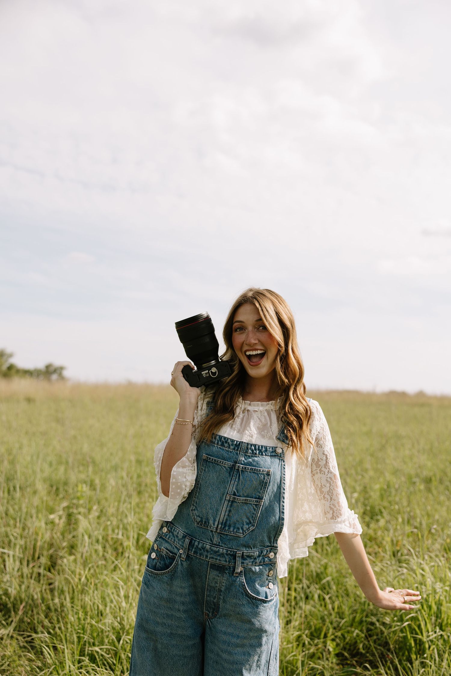 Smiling woman in denim overalls holding a camera in a green field.