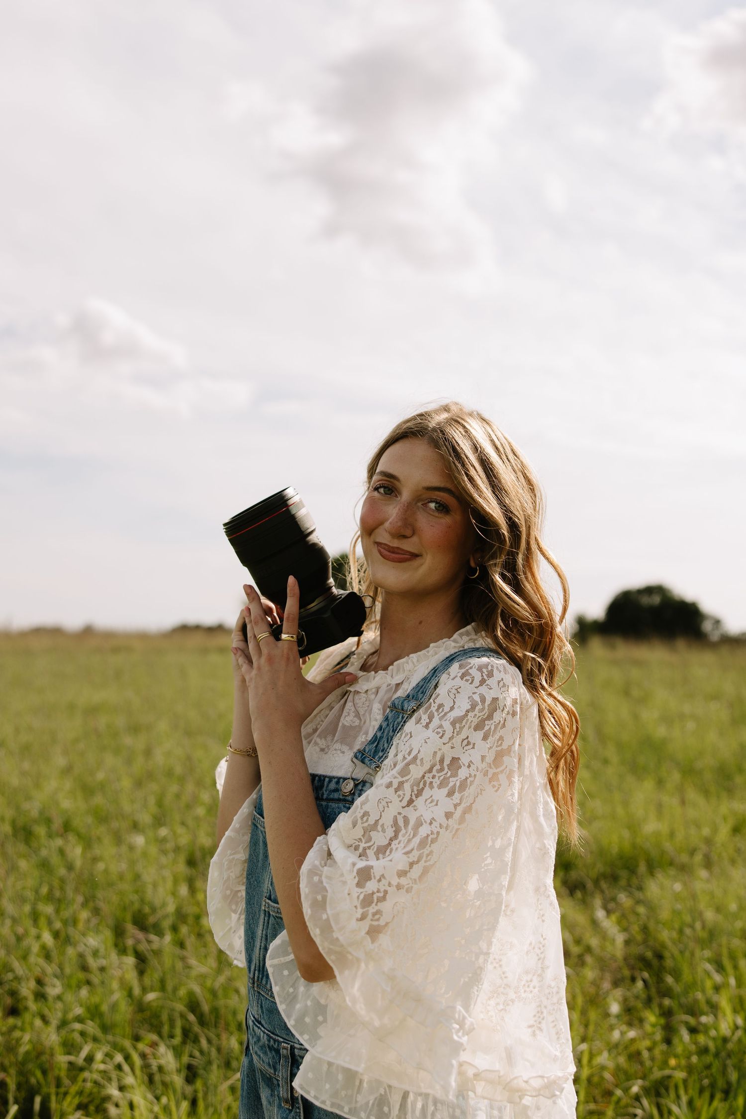 Young woman holding a vintage camera in a green field, wearing lace top and denim overalls, smiling outdoors.