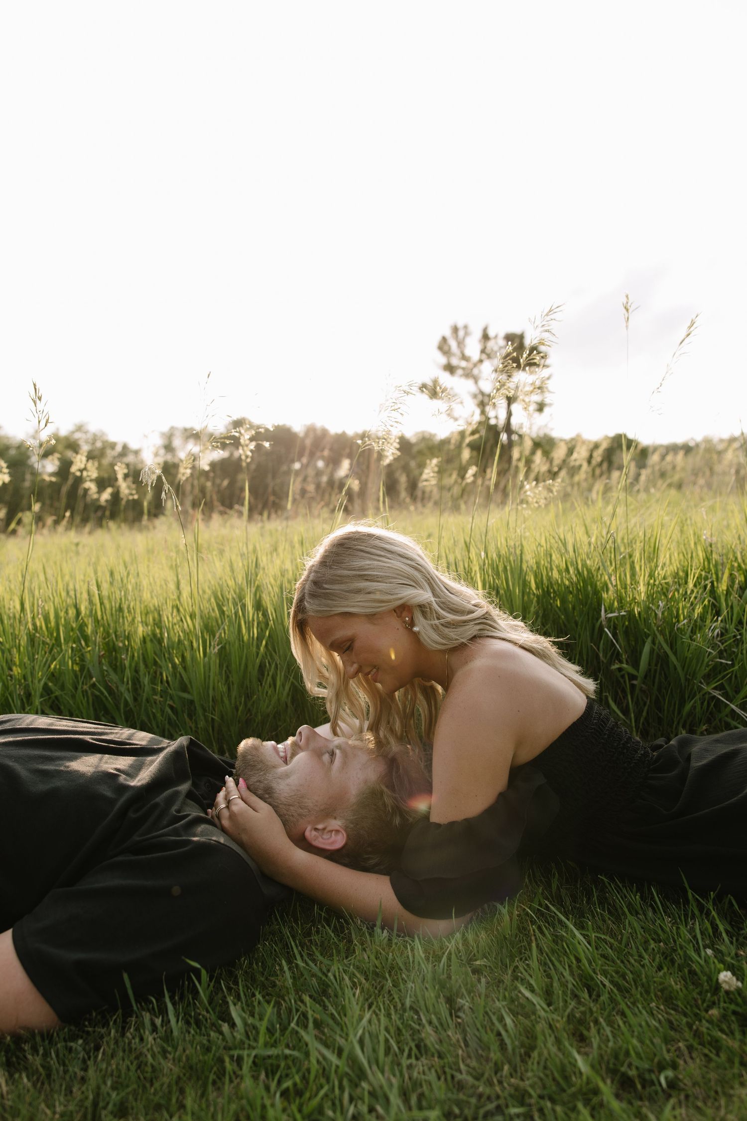 Couple shares a romantic moment in tall green grass field, woman leaning over man lying down at sunset.