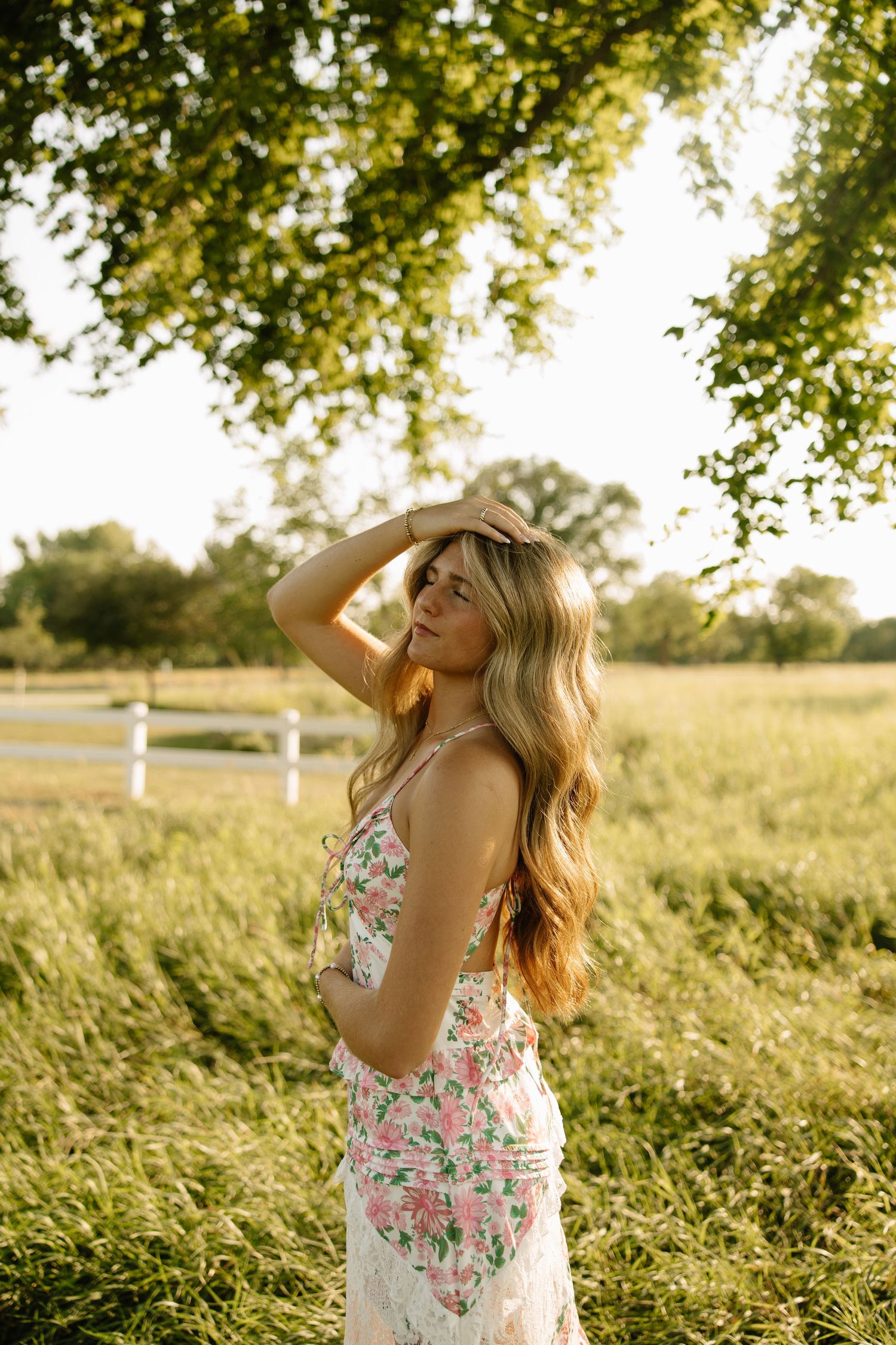 Woman in floral dress poses gracefully under lush green tree in sunlit meadow with white fence backdrop.