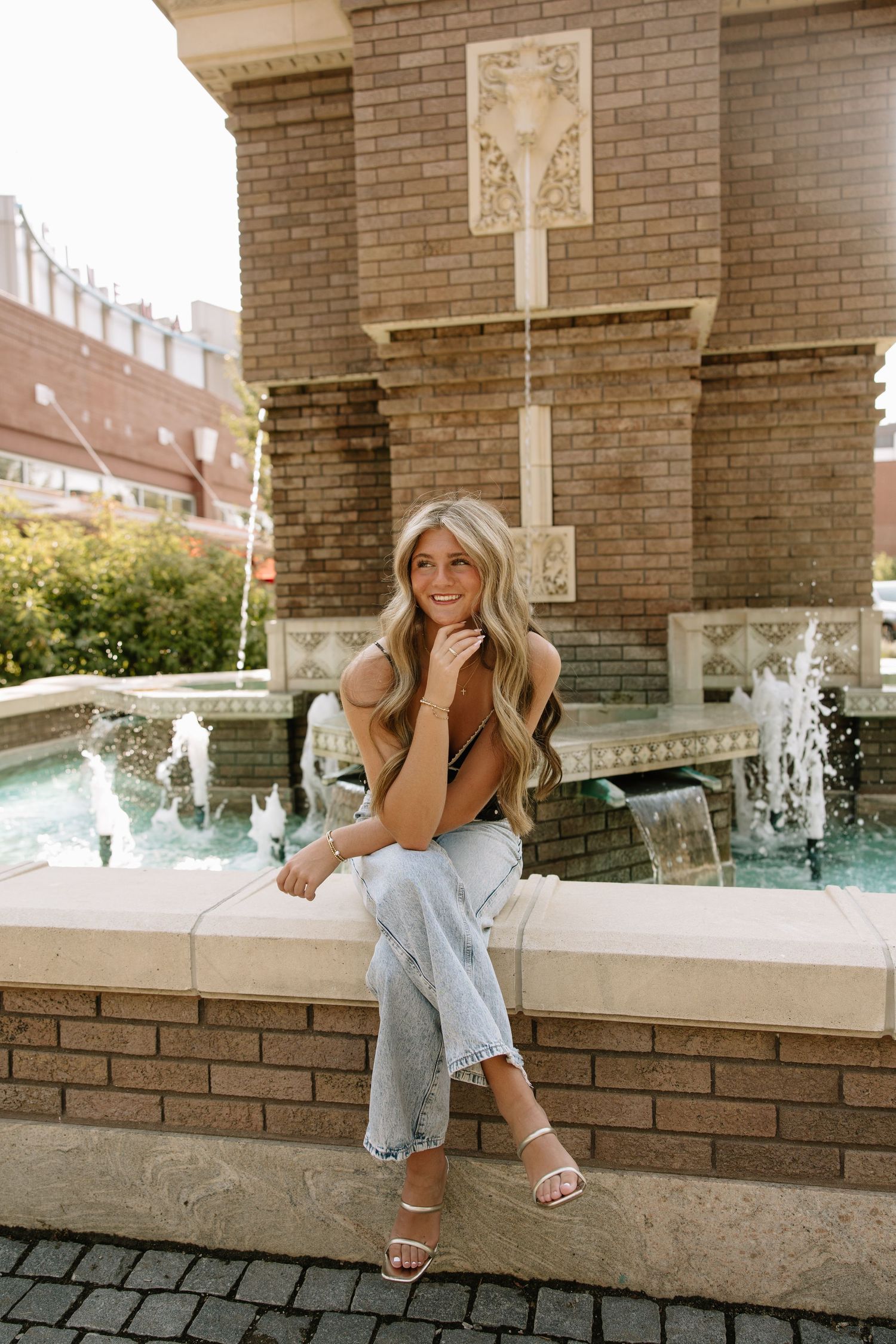 Young woman with long blonde hair sitting on a fountain ledge, wearing jeans and heels, smiling outdoors.