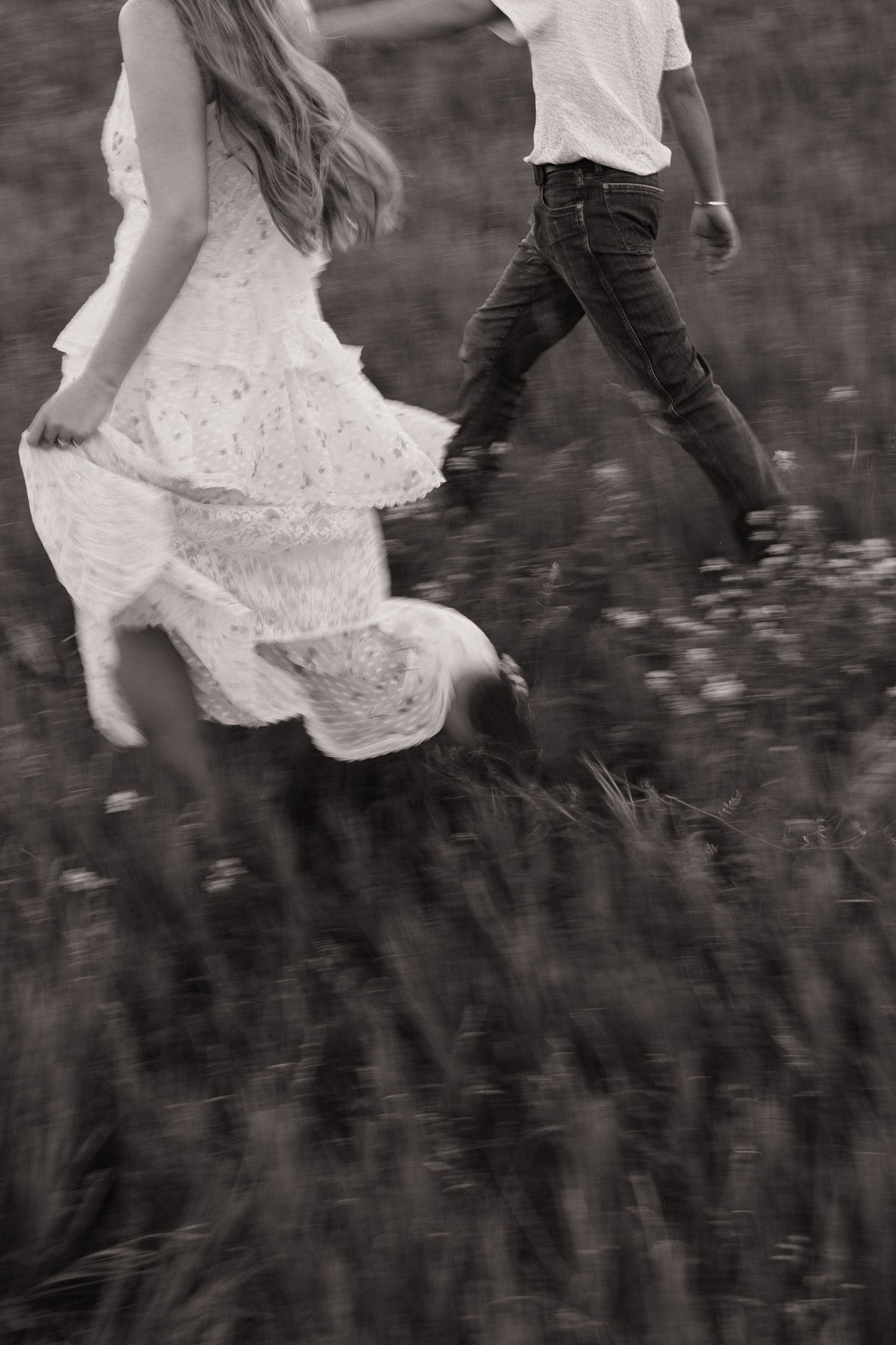 Black and white photo of couple running through wildflower meadow, woman in white dress, man in jeans.