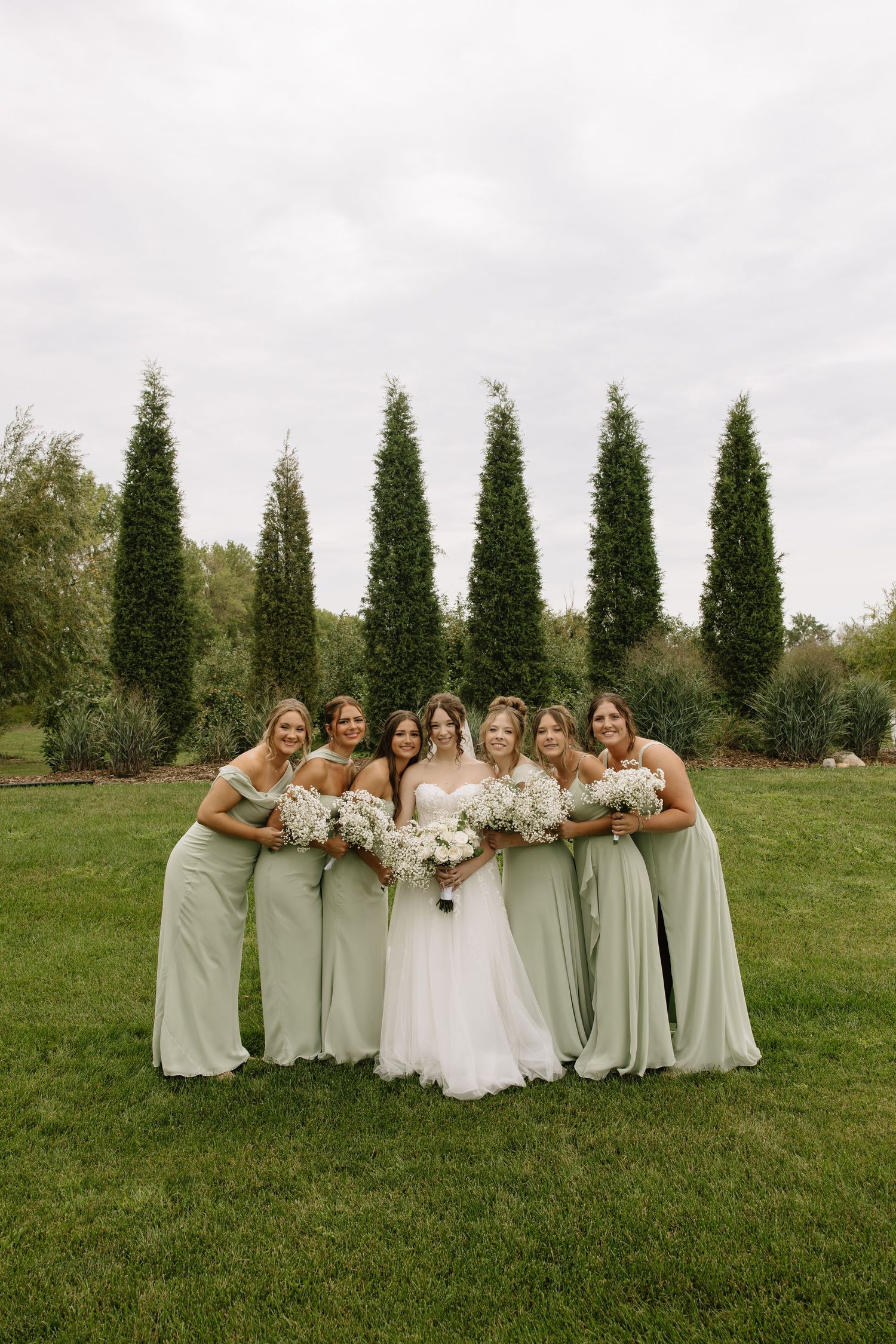 Bride in white gown poses with six bridesmaids in sage green dresses holding white bouquets on a lush green lawn.
