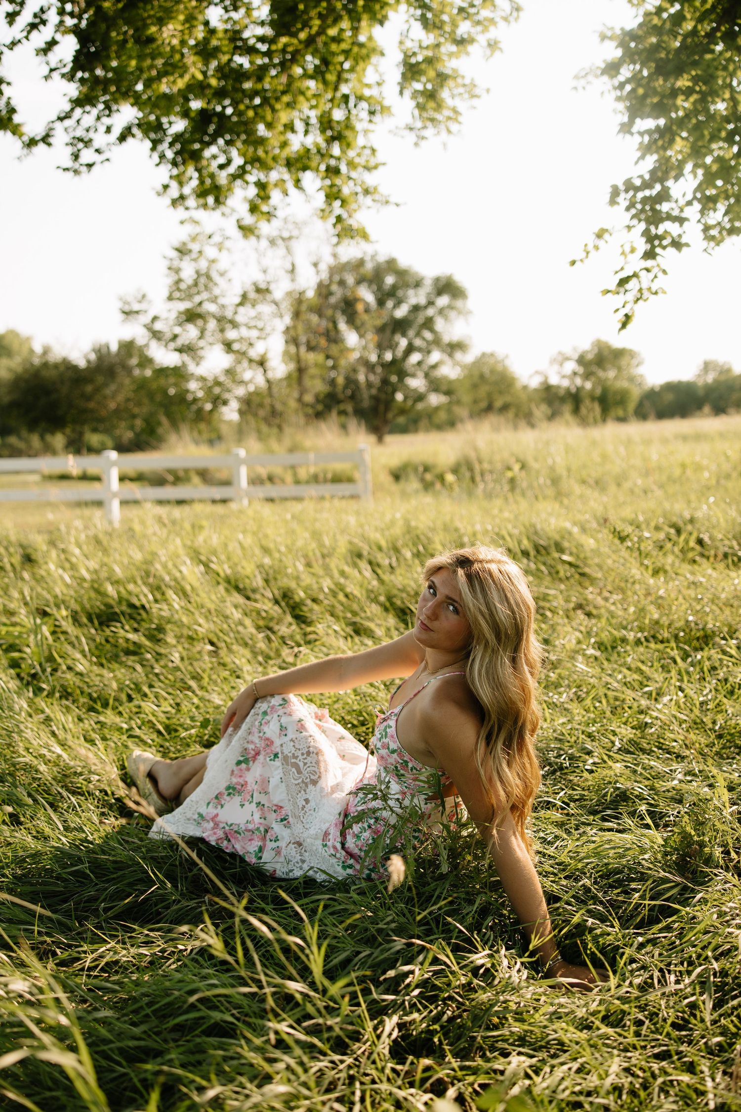 Blonde woman in floral dress sitting in lush green field with white fence and trees in golden sunlight.