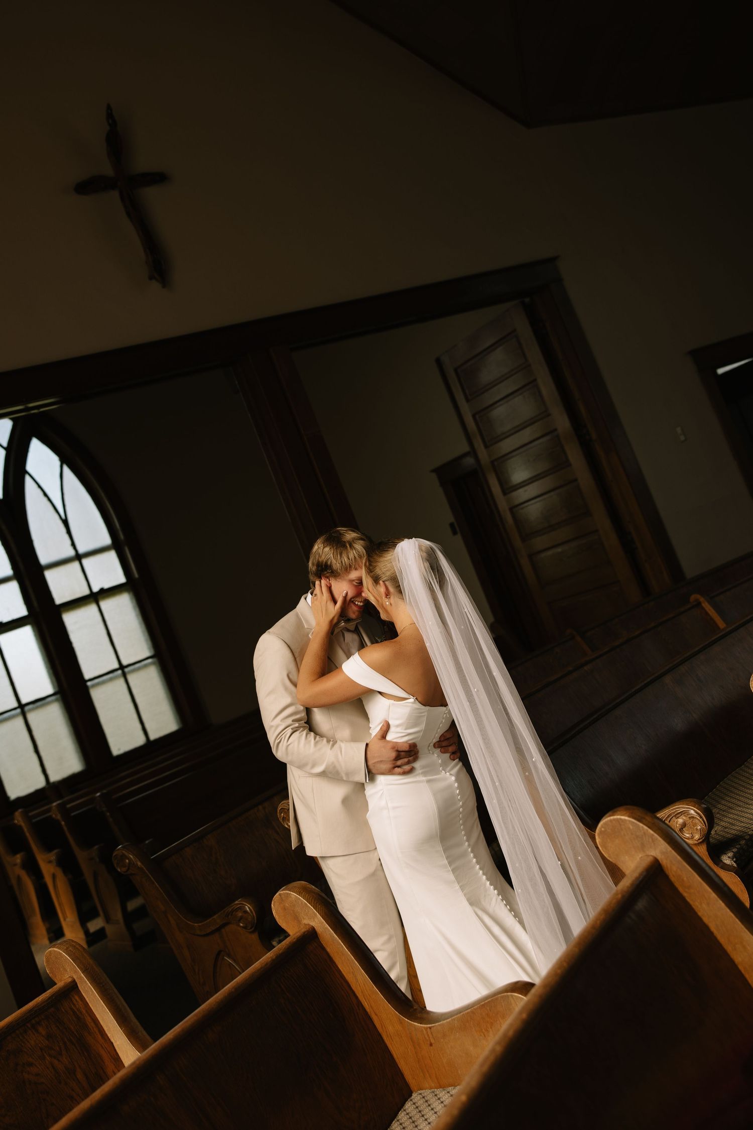 Bride and groom share intimate moment kissing inside dimly lit church with wooden pews and cross on wall.